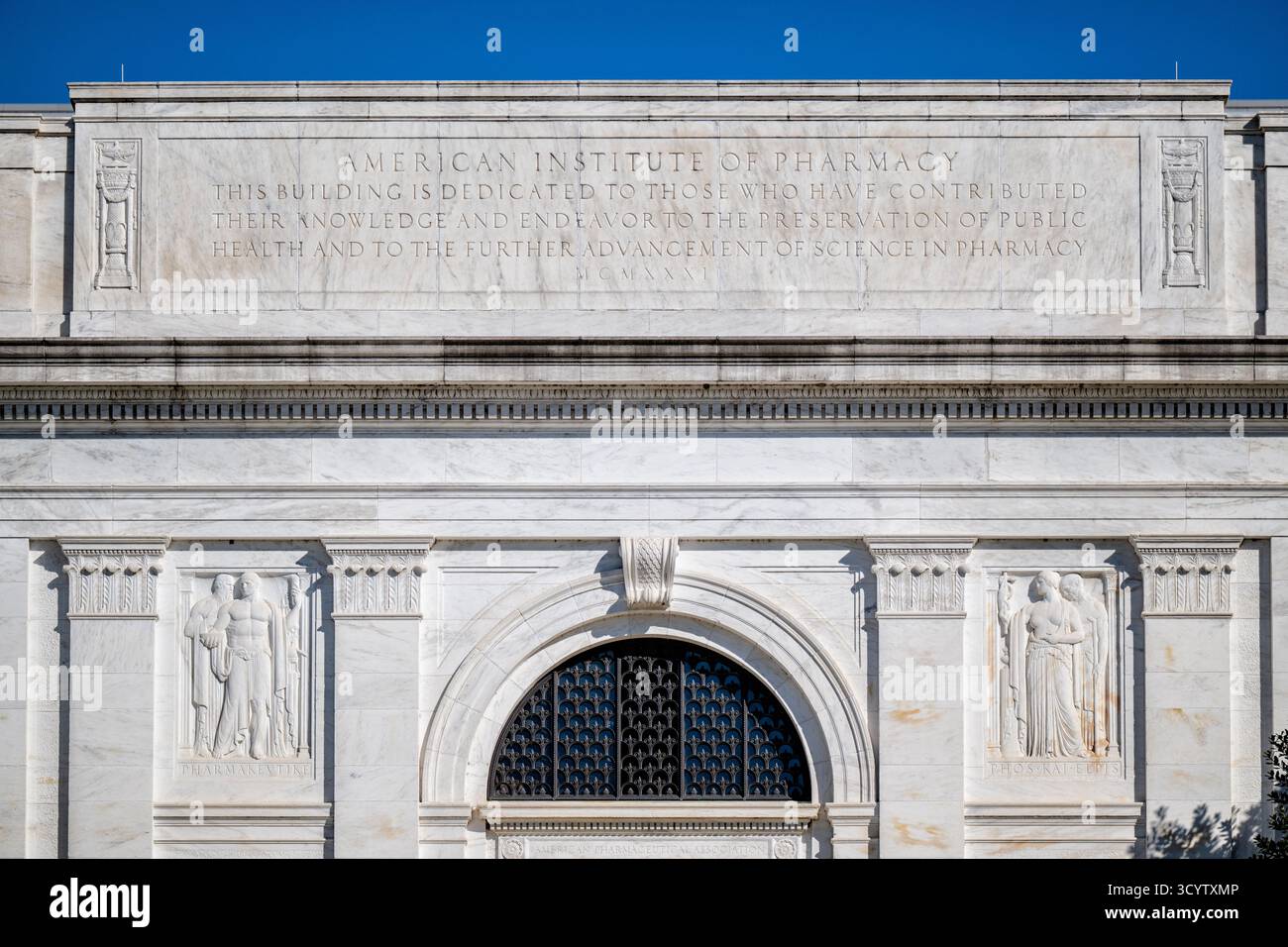 American Institute of Pharmacy Building Pediment Washington DC // WASHINGTON DC — l'American Institute of Pharmacy Building, noto anche come American Pharmaceutical Association Building, presenta un frontone in marmo bianco con un'iscrizione del 1931 (MCMXXXI). Progettata da John Russell Pope, l'iscrizione dedica l'edificio a coloro che hanno avanzato la salute pubblica e la scienza farmaceutica. Sotto, sculture in rilievo fiancheggiano una finestra ad arco, con incisioni 'PHARMAKEVTIKE' (Farmacia) e 'PHOS KAI ELPIS' (luce e speranza). Foto Stock