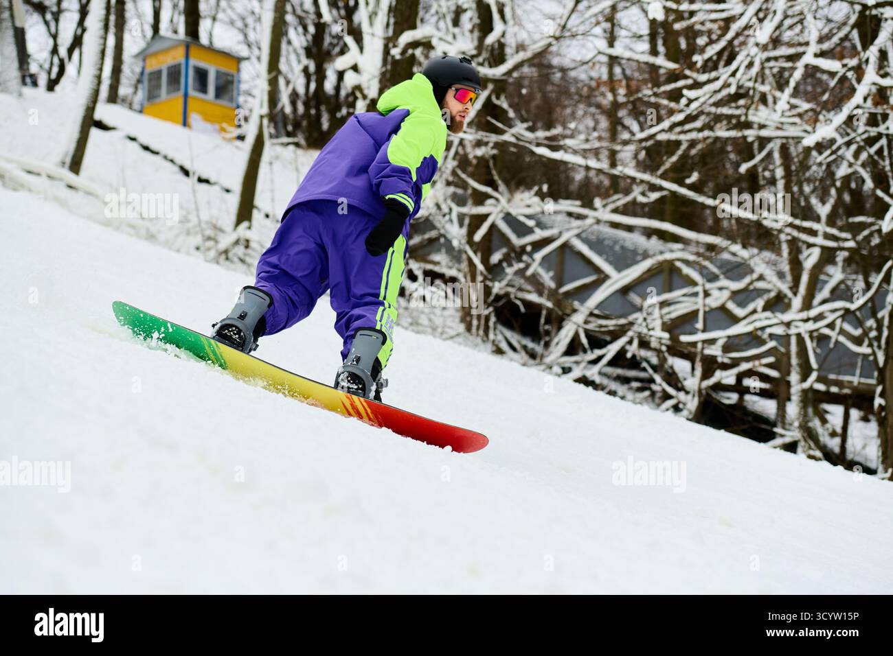 Un uomo barbuto sapientemente fa snowboard su una pista innevata, mostrando vivaci attrezzature invernali. Foto Stock