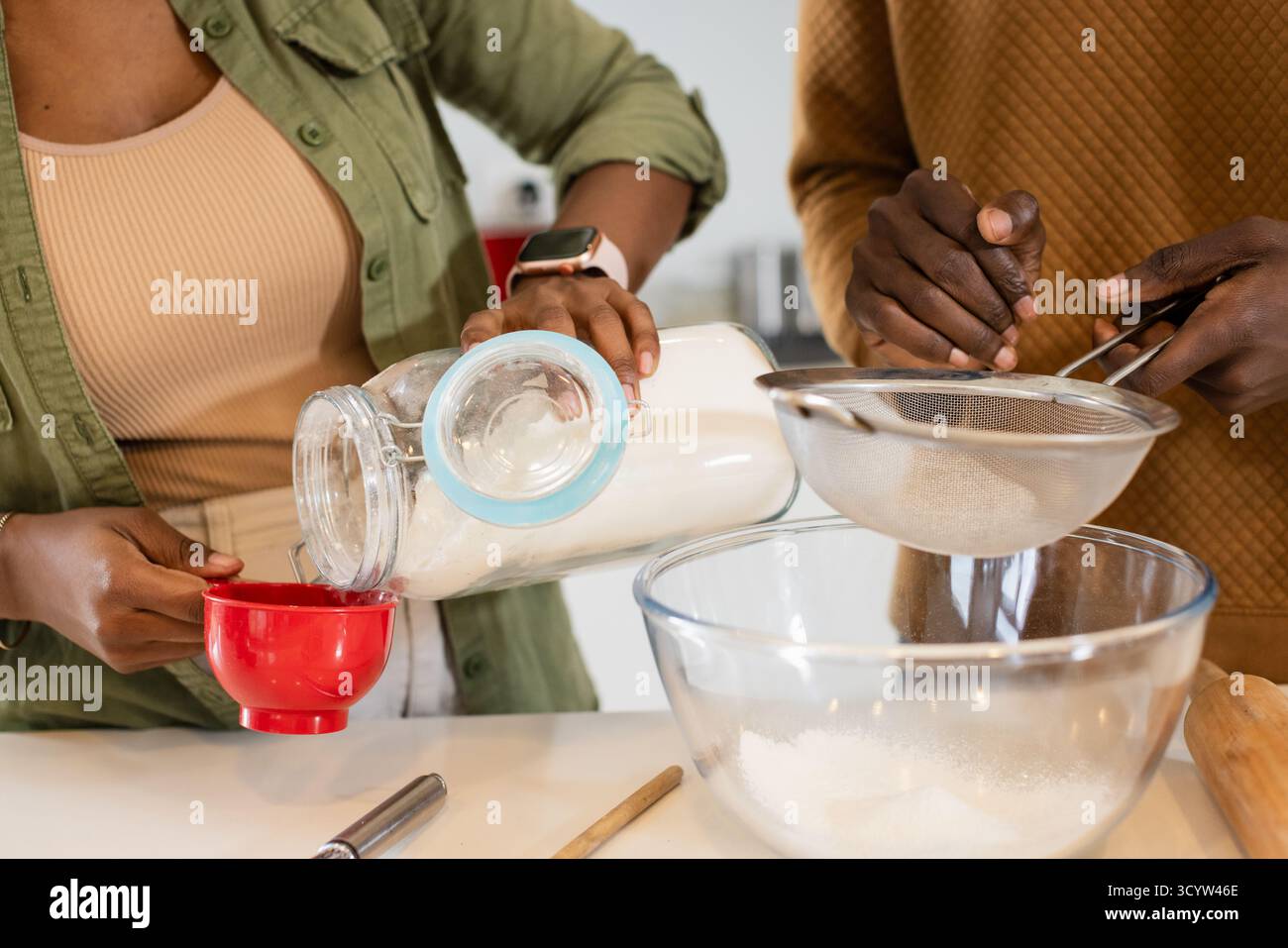 Coppia nera in cucina versando il vaso di farina nella tazza e setacciando il setaccio sopra il recipiente Foto Stock