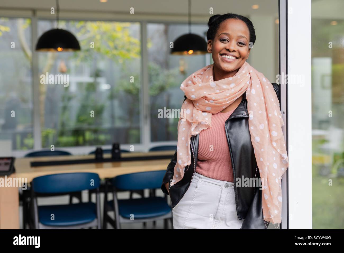 Donna afroamericana appoggiata alla porta, sorridente in ufficio con tavolo, sedie, lampade, spazio fotocopie Foto Stock