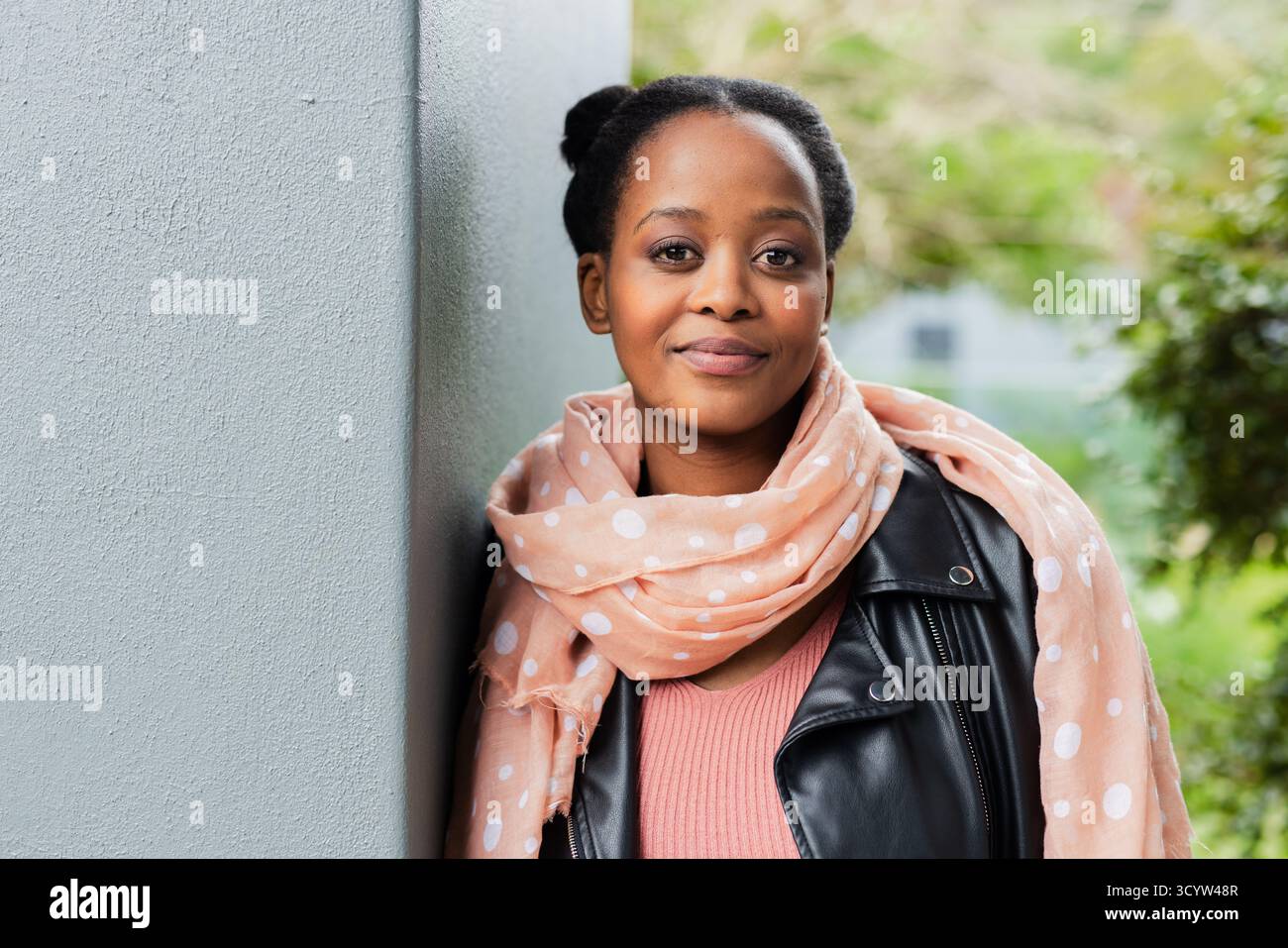 Donna afroamericana all'aperto, accanto alla parete grigia, con giacca in pelle e sciarpa di pesca Foto Stock