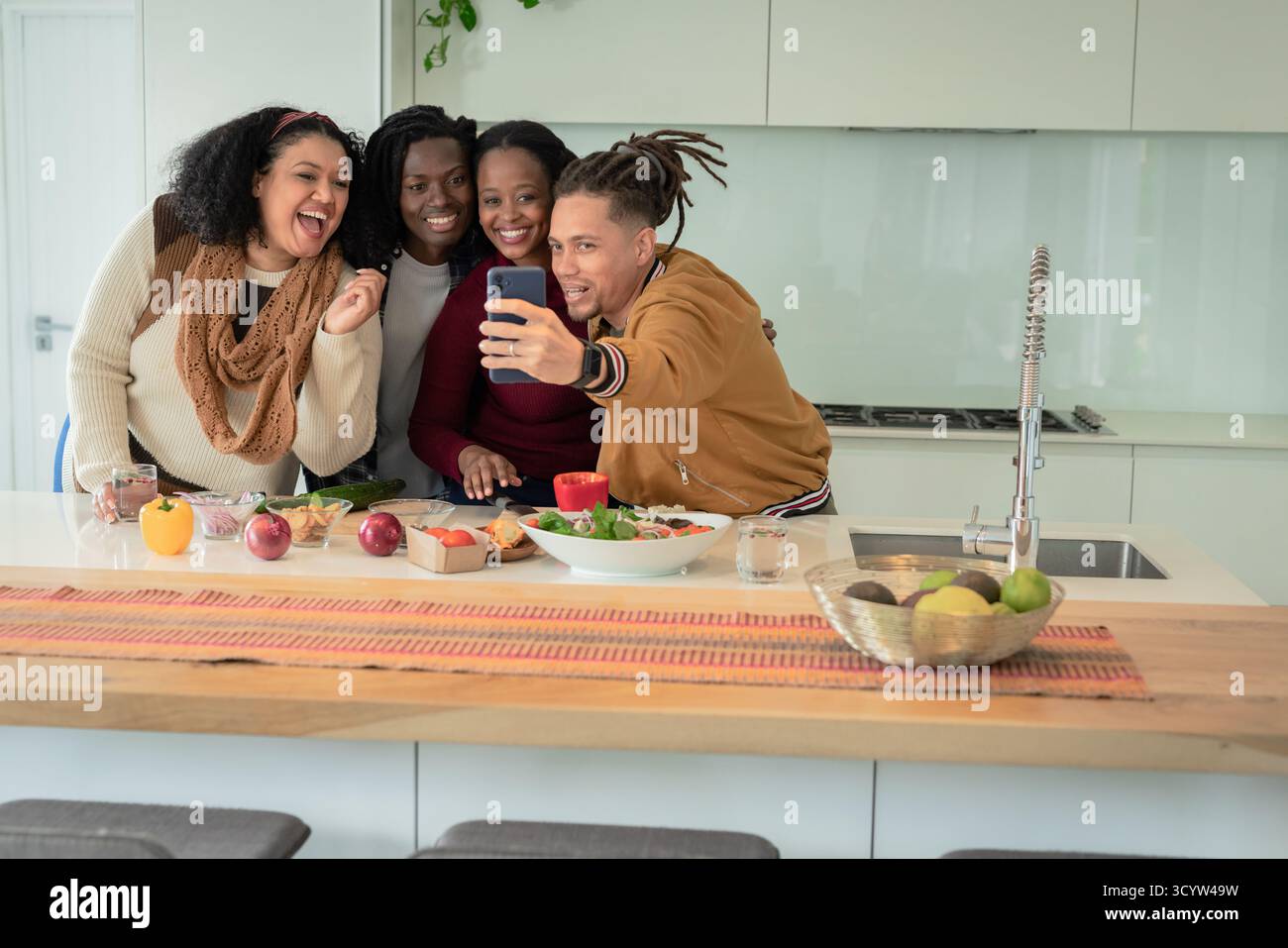 Amici diversi che scattano selfie all'isola della cucina di casa con insalata frutta e smartphone Foto Stock