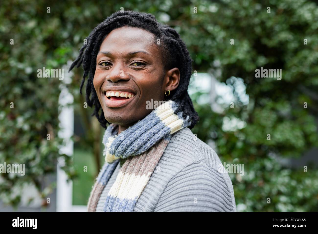 Uomo afro-americano sorridente mentre è in piedi in giardino con maglione grigio e sciarpa a righe. Ritratto, casual, bohémien, stile di vita, natura, rilassato, fuori Foto Stock
