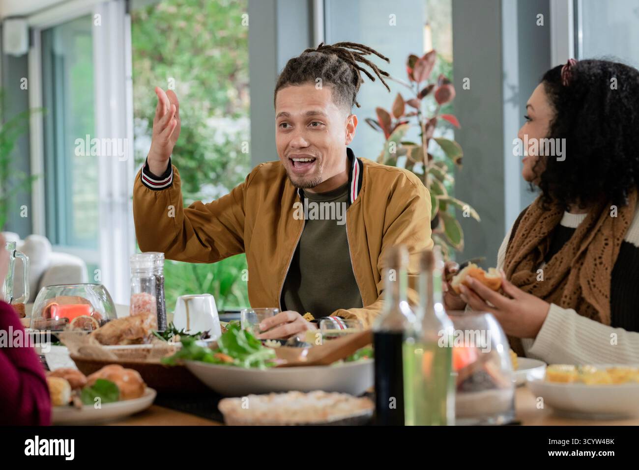 Diversi amici condividono il pranzo nella sala da pranzo di casa intorno a un tavolo di legno con insalata e pasticcini Foto Stock