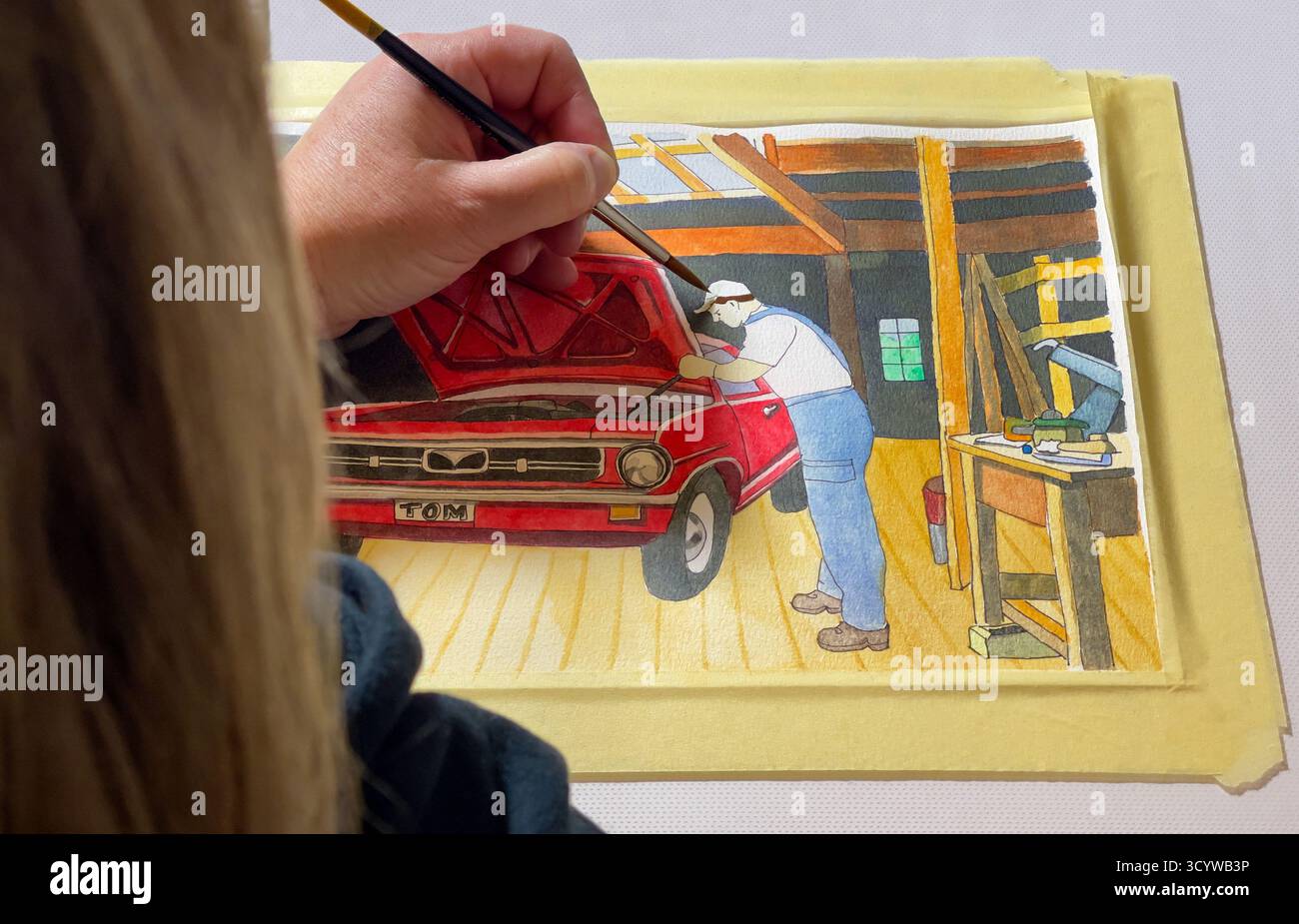 Una donna bionda dipinge un acquerello con la mano sinistra. L'acquerello mostra un meccanico di auto in piedi accanto al cofano aperto di un'auto d'epoca rossa. L'auto lo è Foto Stock