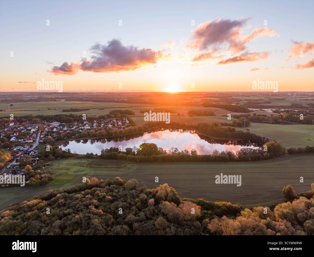 il lago still rispecchia il cielo scintillante del tramonto, circondato da alberi autunnali, campi aperti e un tranquillo villaggio immerso nel tranquillo paesaggio rurale Foto Stock