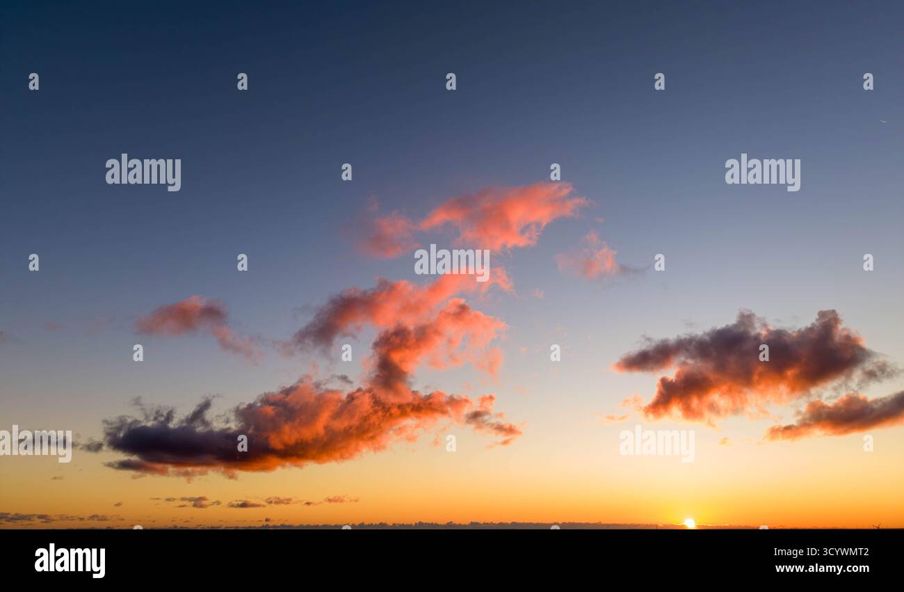 scena del tramonto con nuvole arancioni e rosa sparse in un cielo limpido mentre il sole scende sotto l'orizzonte, emettendo un bagliore caldo e sereno Foto Stock