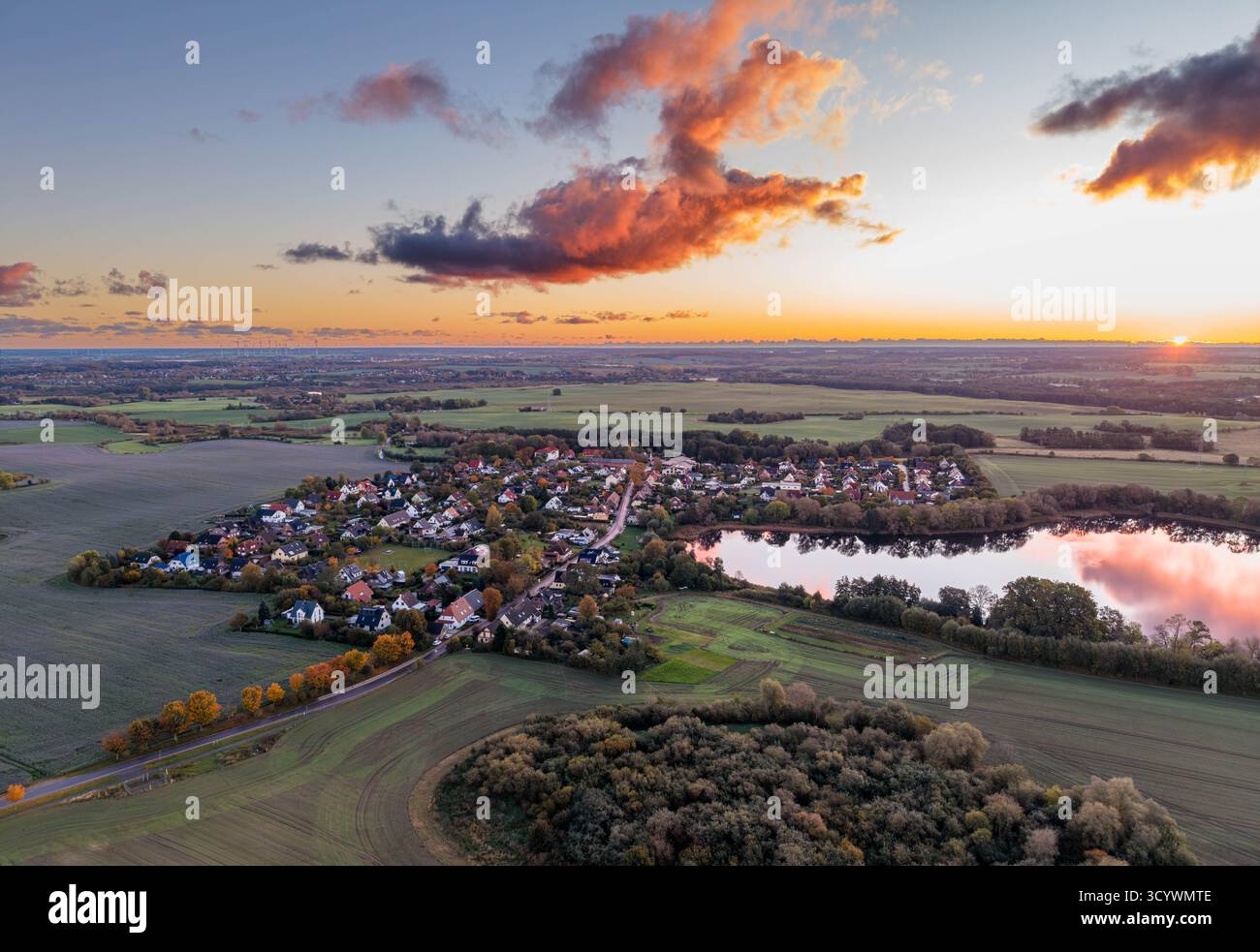 tranquillo villaggio di campagna è annidato tra campi e foreste, con un lago riflettente nelle vicinanze, il tutto immerso nella luce soffusa e sbiadita di un colorato su Foto Stock