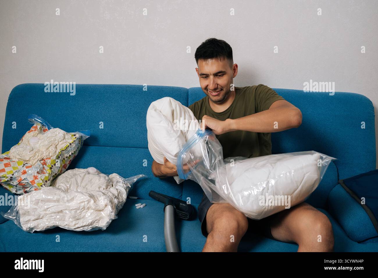 Un giovane sorridente che confeziona cuscini bianchi e biancheria da letto in sacchetti sottovuoto seduti sul divano blu del soggiorno, pronti a muoversi o a riordinare Foto Stock