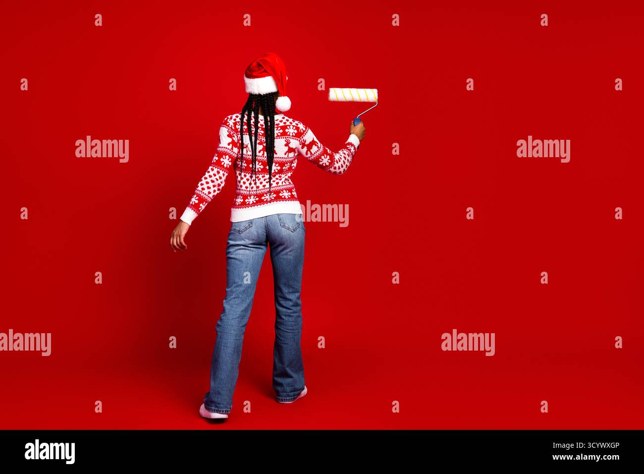 Una divertente pittrice delle feste con un maglione natalizio dipinge una parete rossa con un rullo mentre una donna sorridente indossa il cappello e i jeans di Babbo Natale Foto Stock
