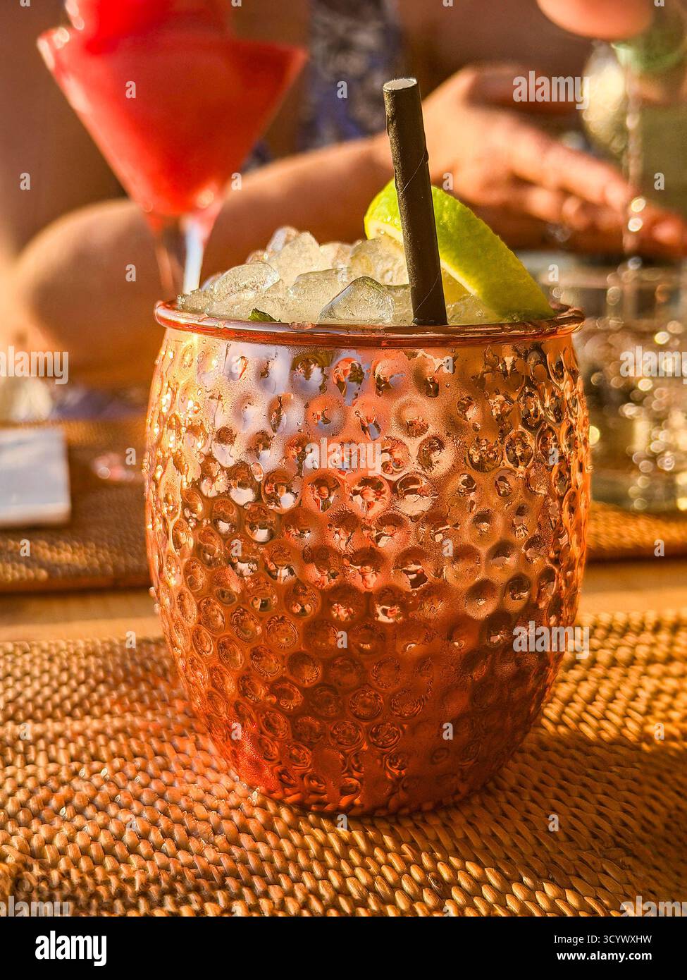 Cocktail mulo moscovita in una tazza di rame martellata con fetta di lime e paglia su un tappeto intrecciato, con persone in sottofondo che si godono delle bevande Foto Stock