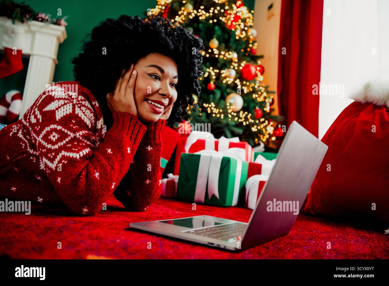 La donna sorridente in maglione rosso ama le videochiamate durante le festività natalizie vicino a alberi decorati e regali luminosi. Foto Stock