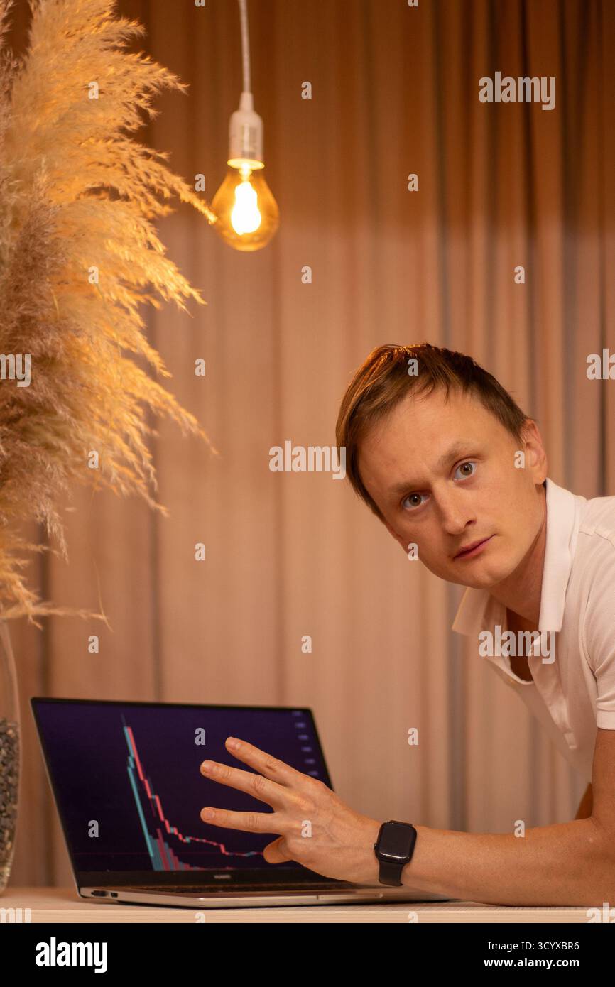 Giovane attento e teso che analizza grafici del mercato delle criptovalute e dati di trading su notebook nel moderno spazio di lavoro per gli uffici domestici. Affari, amico. Foto Stock