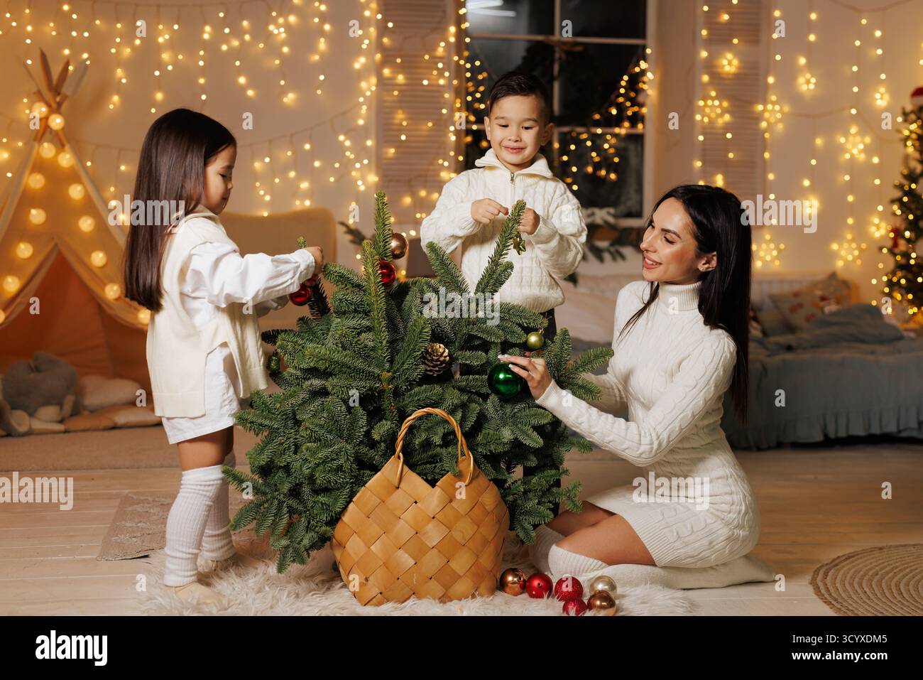 Mamma e bambini decorano un piccolo abete rosso in una casa accogliente con luci scintillanti, ideale per campagne di vacanza, biglietti e annunci stagionali. Foto Stock