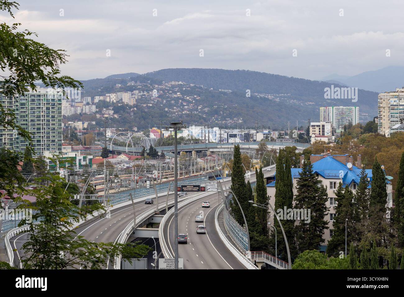 Ampia autostrada sul fiume urbano. Ampia autostrada sopraelevata che attraversa un fiume in una città vivace con edifici alti, viali cipressi, tetti blu, e. Foto Stock