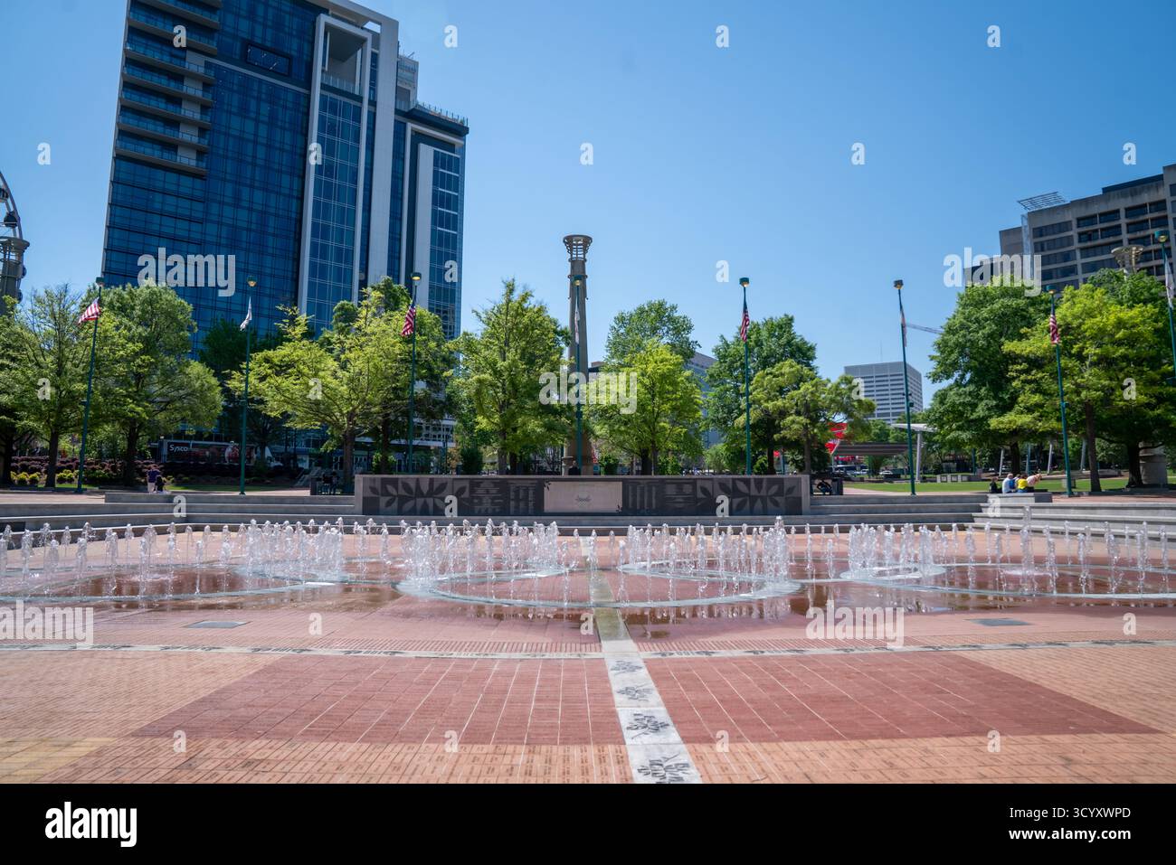 I getti d'acqua sorgono dalla fontana degli anelli Olimpici del Centennial Olympic Park, incorniciati da moderni grattacieli, alberi e bandiere americane sotto un cielo limpido Foto Stock