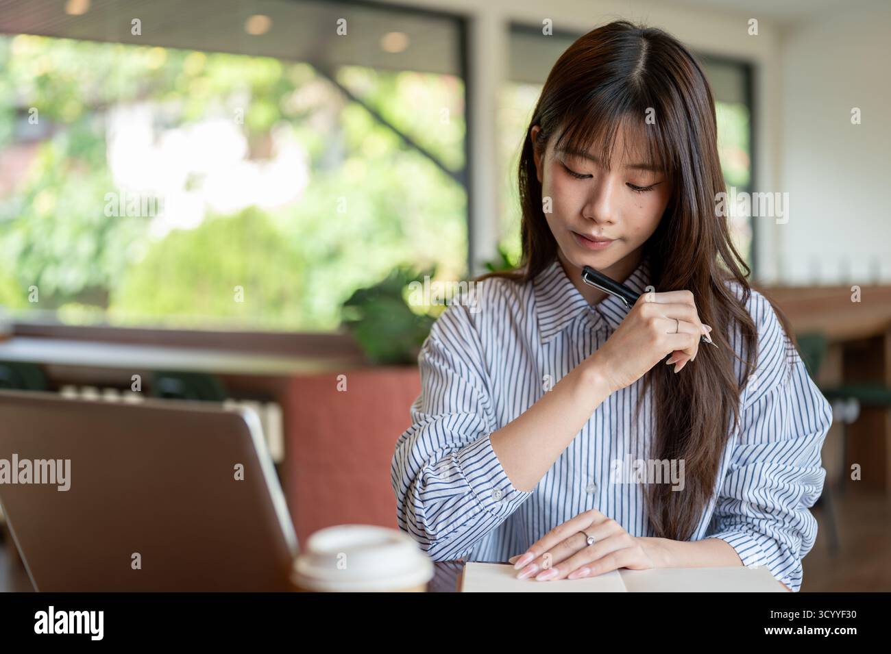 Donna asiatica in camicia a righe blu che tiene una stilografica e guarda il notebook mentre siede al tavolo nel bar. Studiare o lavorare all'esterno. Foto Stock