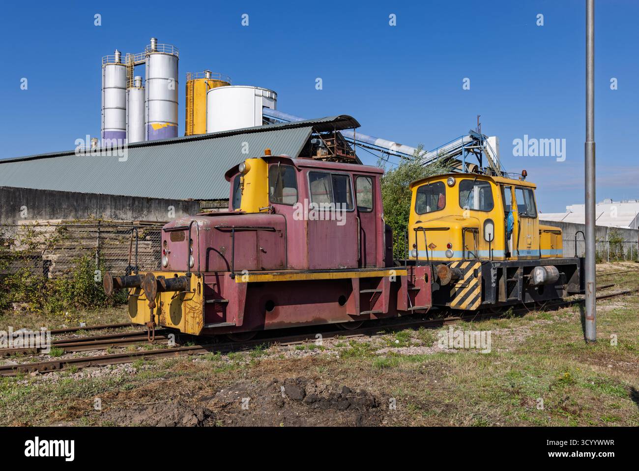 Due locomotive industriali abbandonate, una gialla e una viola/rossa, situate su binari ferroviari vicino a un impianto industriale di cemento di Basal. Foto Stock