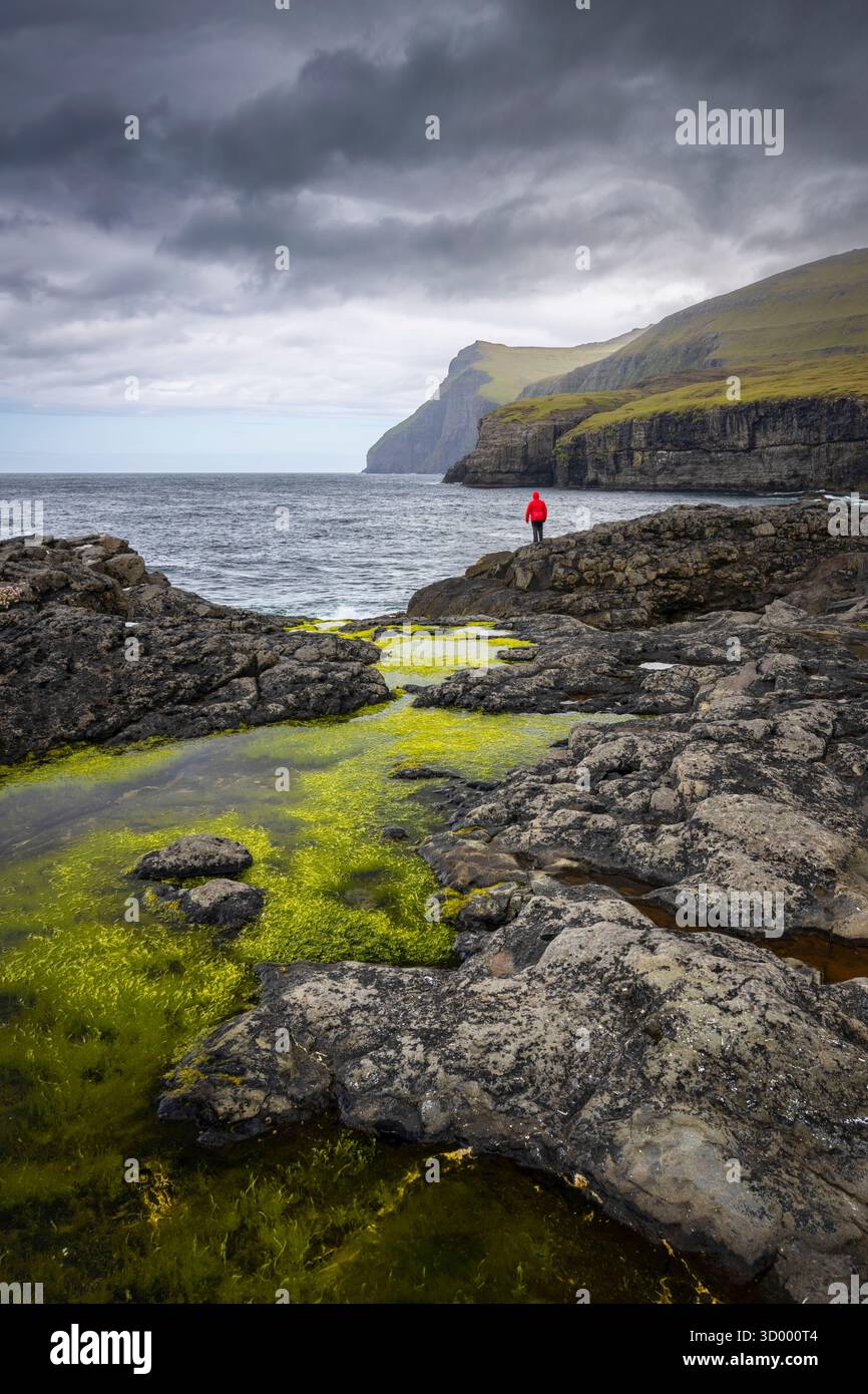 Vista di una persona vestita di rosso che ammira la costa selvaggia vicino al villaggio di Eidi. Isola di Eysturoy, Isole Faroe, Danimarca, Europa. Foto Stock