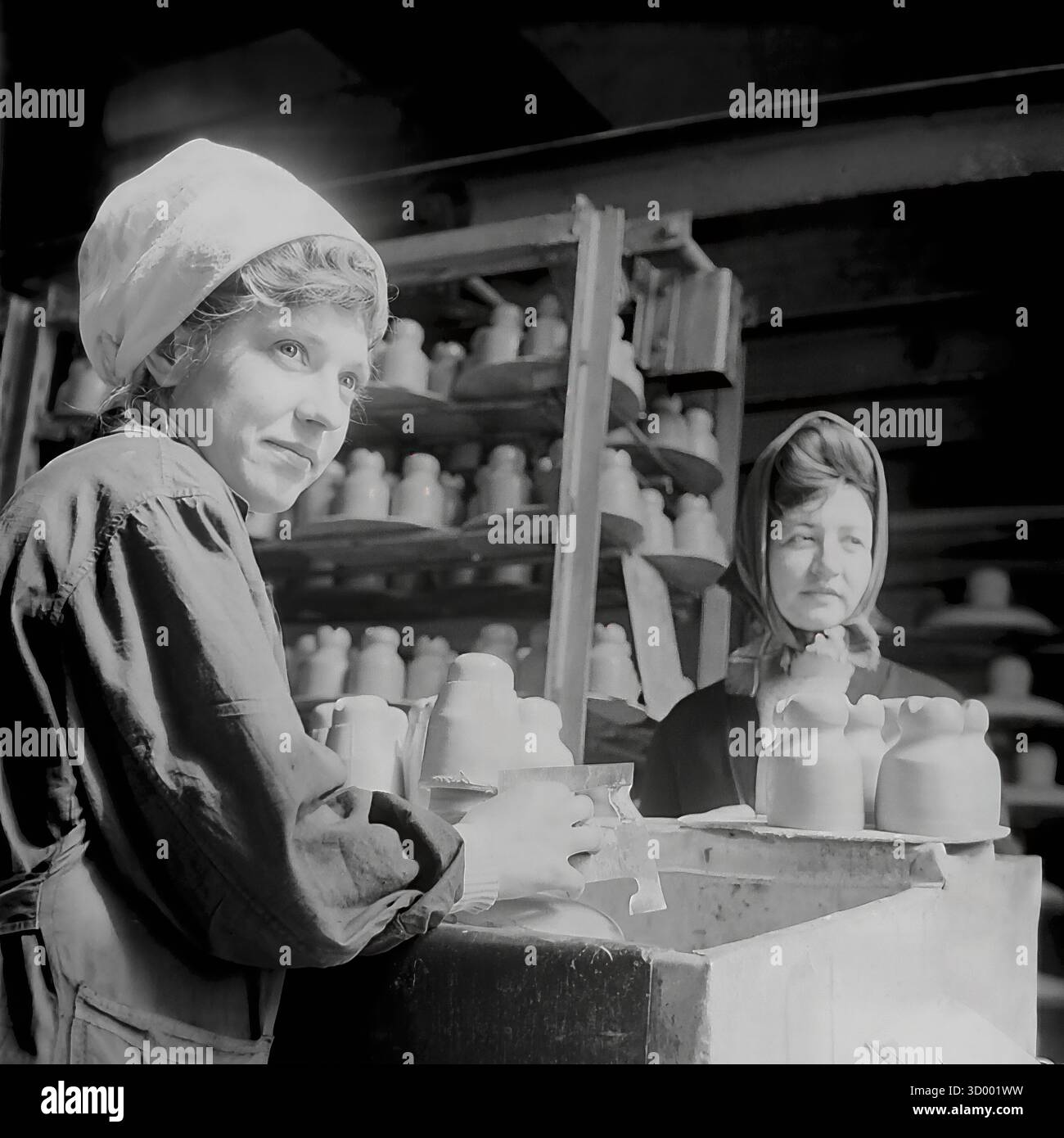 Questa foto d'archivio del 1966 di Sloviansk, RSS Ucraina, cattura un momento sincero presso la fabbrica di armature e isolatori (AIF). Presenta una giovane lavoratrice alla sua postazione, che indossa una copertura della testa di colore chiaro e una scrofetta da lavoro. Sta maneggiando isolanti di argilla bagnati. Guarda in alto, sorride. Un'altra collega con un velo più scuro si trova nelle vicinanze. Lo sfondo è pieno di rack impilati in alto con prodotti in ceramica. Questo ritratto mette in evidenza l'elemento umano e il ruolo delle donne nell'industria pesante sovietica, un documento del pacifico Donbas. Foto Stock