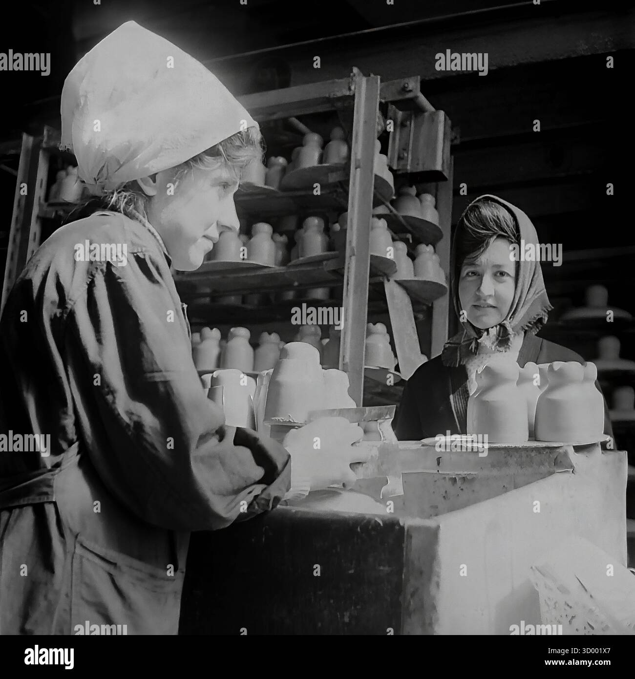 Questa foto d'archivio del 1966 di Sloviansk, RSS Ucraina, cattura un momento sincero presso la fabbrica di armature e isolatori (AIF). Presenta una giovane lavoratrice alla sua postazione, che indossa una copertura della testa di colore chiaro e una scrofetta da lavoro. Sta maneggiando isolanti di argilla bagnati. Guarda in alto, sorride. Un'altra collega con un velo più scuro si trova nelle vicinanze. Lo sfondo è pieno di rack impilati in alto con prodotti in ceramica. Questo ritratto mette in evidenza l'elemento umano e il ruolo delle donne nell'industria pesante sovietica, un documento del pacifico Donbas. Foto Stock