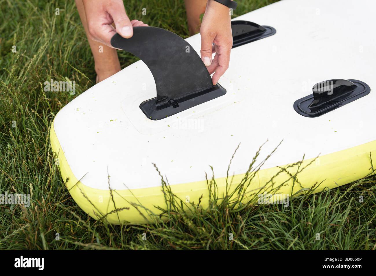 L'immagine mostra un uomo che attacca una pinna nera sul fondo di una tavola da surf bianca e gialla posizionata su un'erba verde lussureggiante. Lezioni di surf Foto Stock
