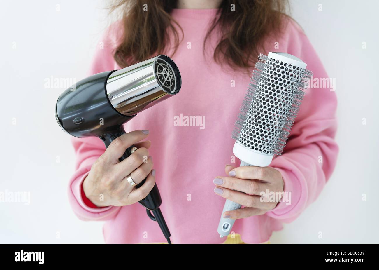 La ragazza tiene in mano un asciugacapelli e una spazzola. Asciugatura dei capelli castani lunghi con una spazzola rotonda per acconciare i capelli senza scrupoli. Salone di bellezza Foto Stock
