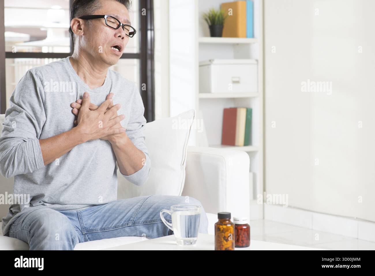 Uomo asiatico maturo degli anni '50, che preme sul petto con espressione dolorosa, seduto sul divano a casa, medicine sul tavolo Foto Stock