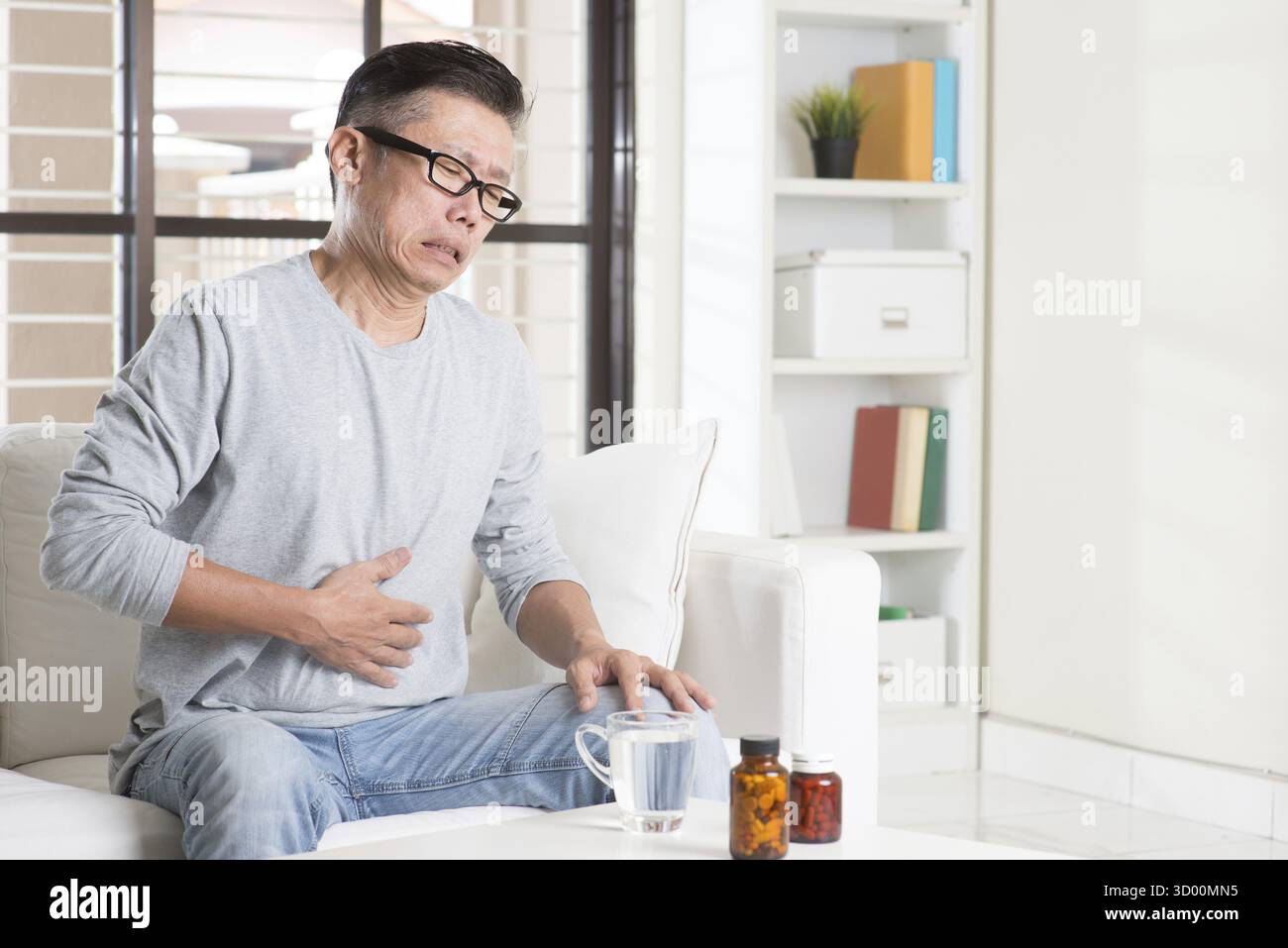 Uomo asiatico maturo anni '50 mal di stomaco, pressare lo stomaco con espressione dolorosa, sedersi sul divano a casa, farmaci sul tavolo Foto Stock