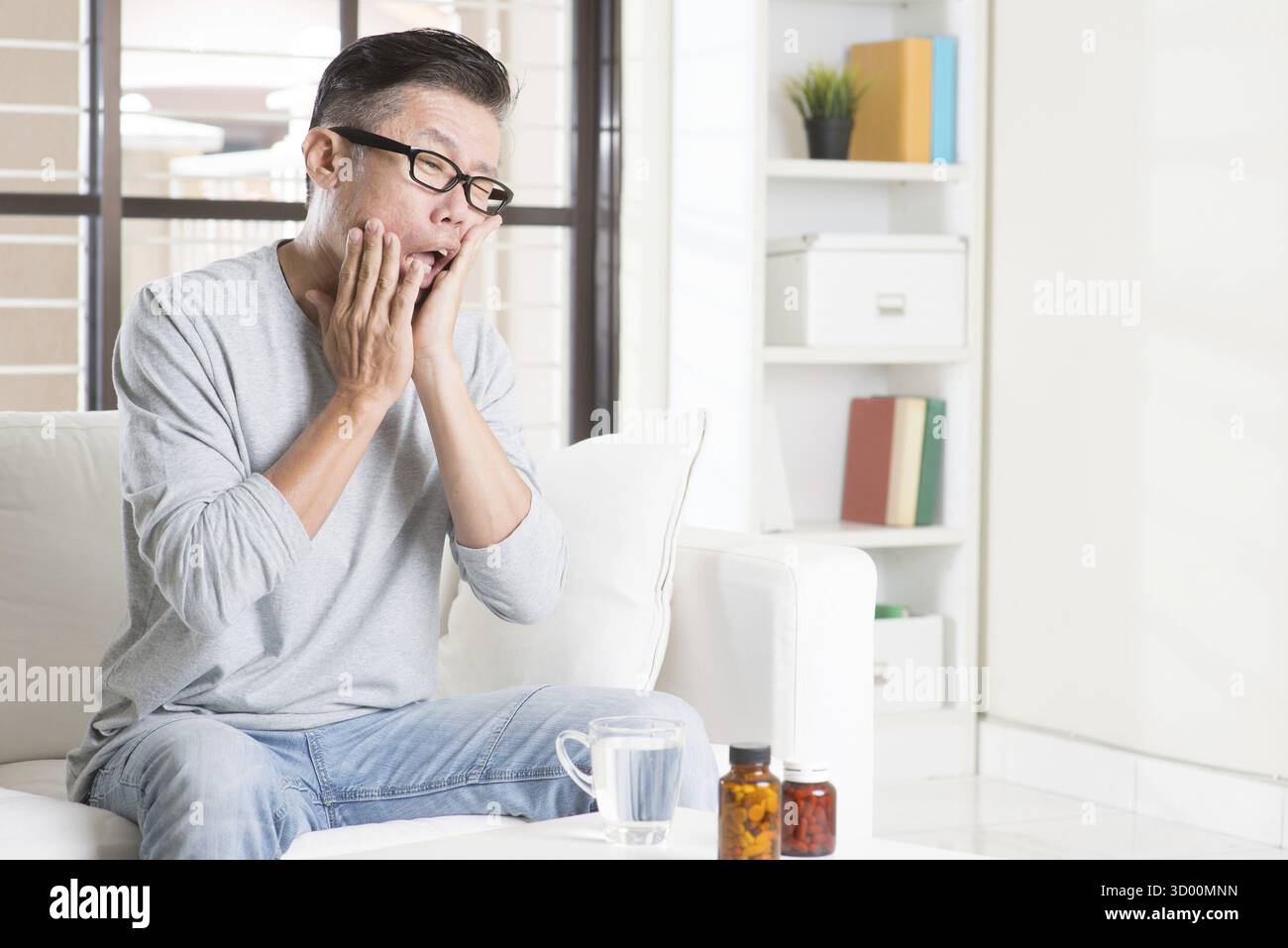 Ritratto di casual 50s coppia uomo asiatico mal di denti con espressione dolorosa, seduto sul divano di casa, medicinali e acqua sul tavolo Foto Stock
