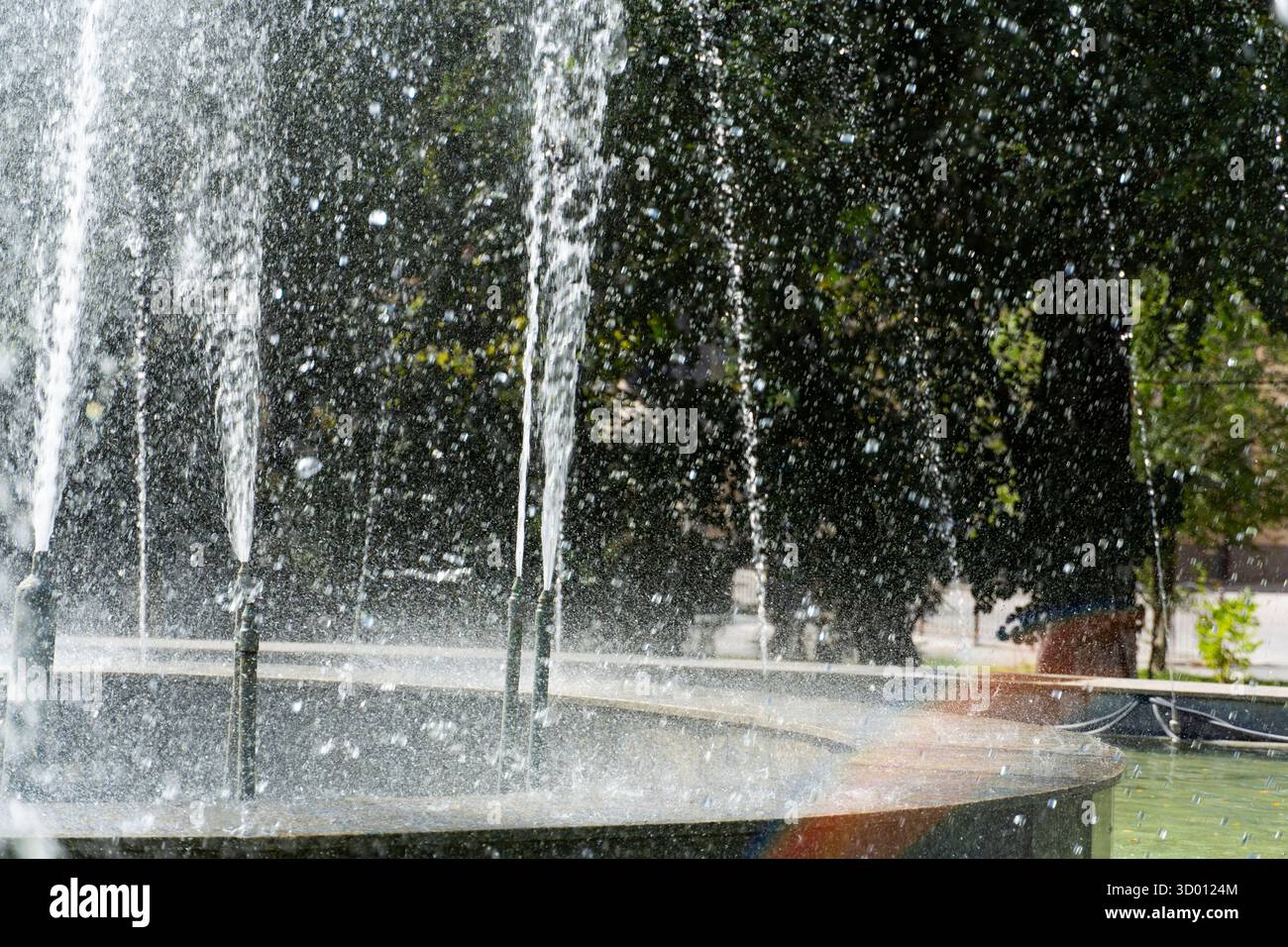 Elegante fontana circolare con alto getto centrale e ruscelli d'acqua circostanti in un parco verde sotto il cielo azzurro, a simboleggiare il relax e la vita urbana Foto Stock
