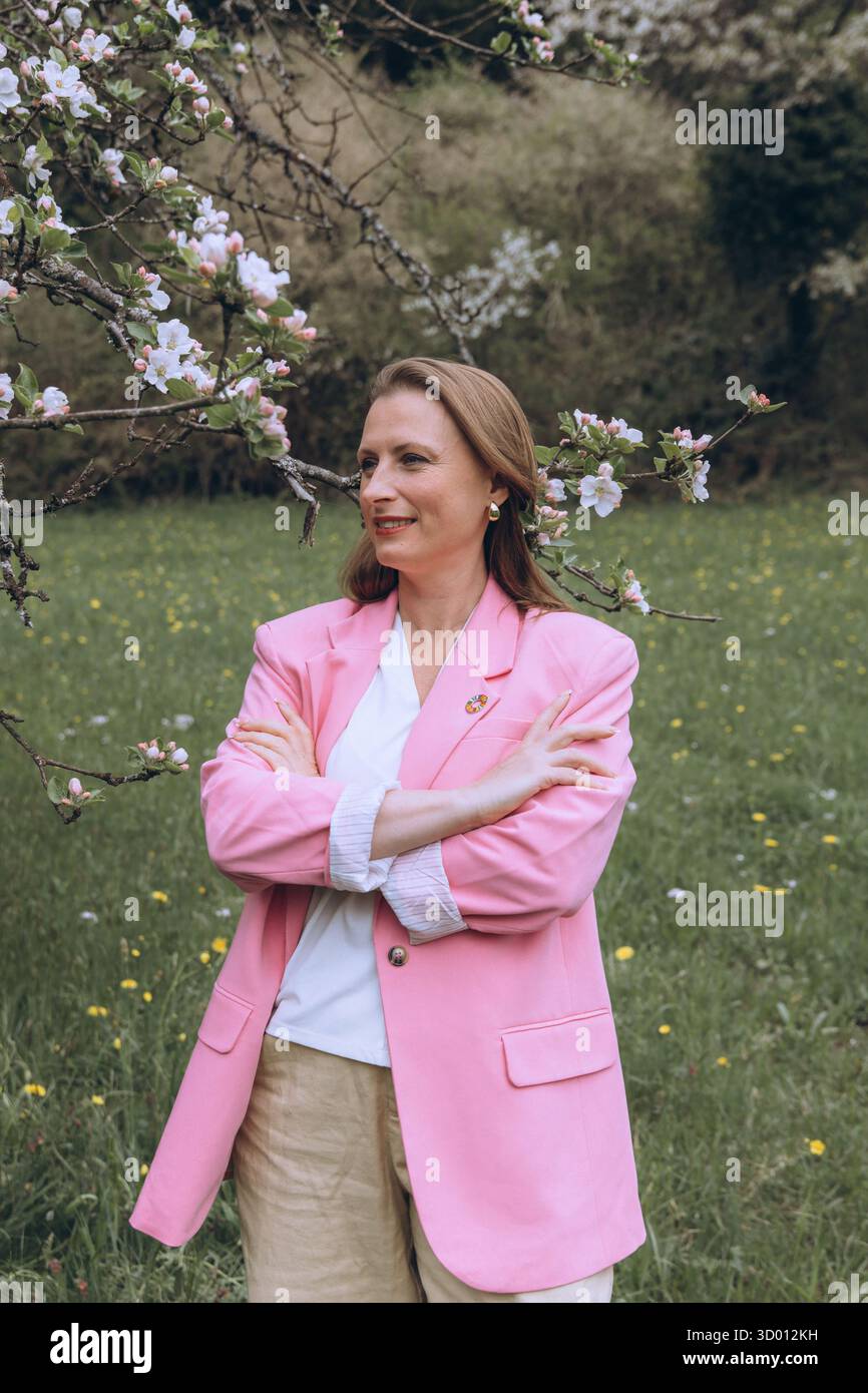 Una donna sorridente di mezza età si trova vicino a un albero in fiore nel giardino primaverile. Bellezza naturale, gioia e armonia con la natura. Rinnovamento stagionale, pace interiore e. Foto Stock