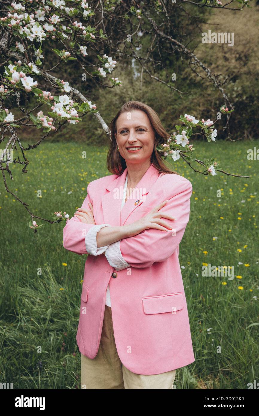 Una donna sorridente di mezza età si trova vicino a un albero in fiore nel giardino primaverile. Bellezza naturale, gioia e armonia con la natura. Rinnovamento stagionale, pace interiore e. Foto Stock
