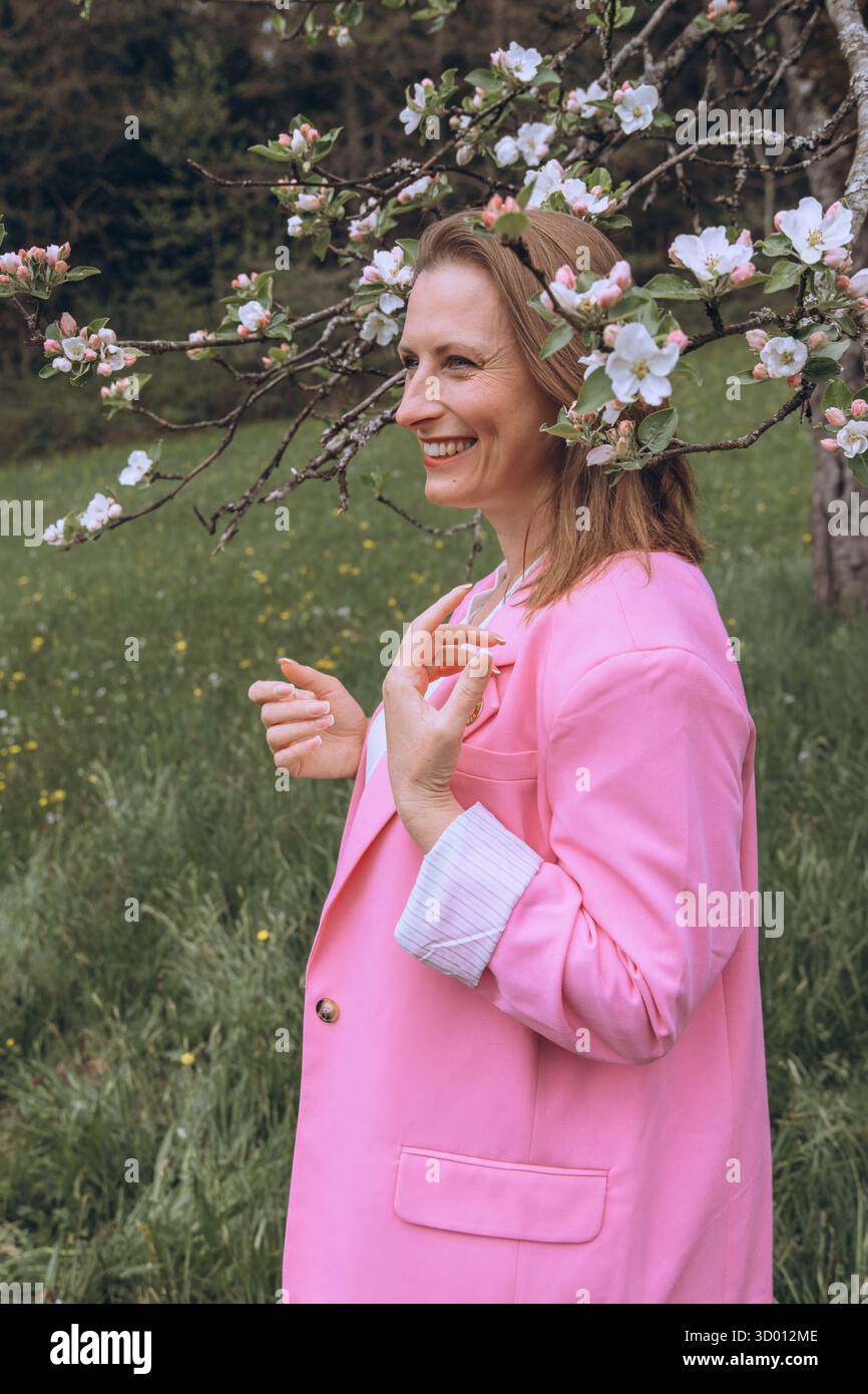 Una donna sorridente di mezza età si trova vicino a un albero in fiore nel giardino primaverile. Bellezza naturale, gioia e armonia con la natura. Rinnovamento stagionale, pace interiore e. Foto Stock
