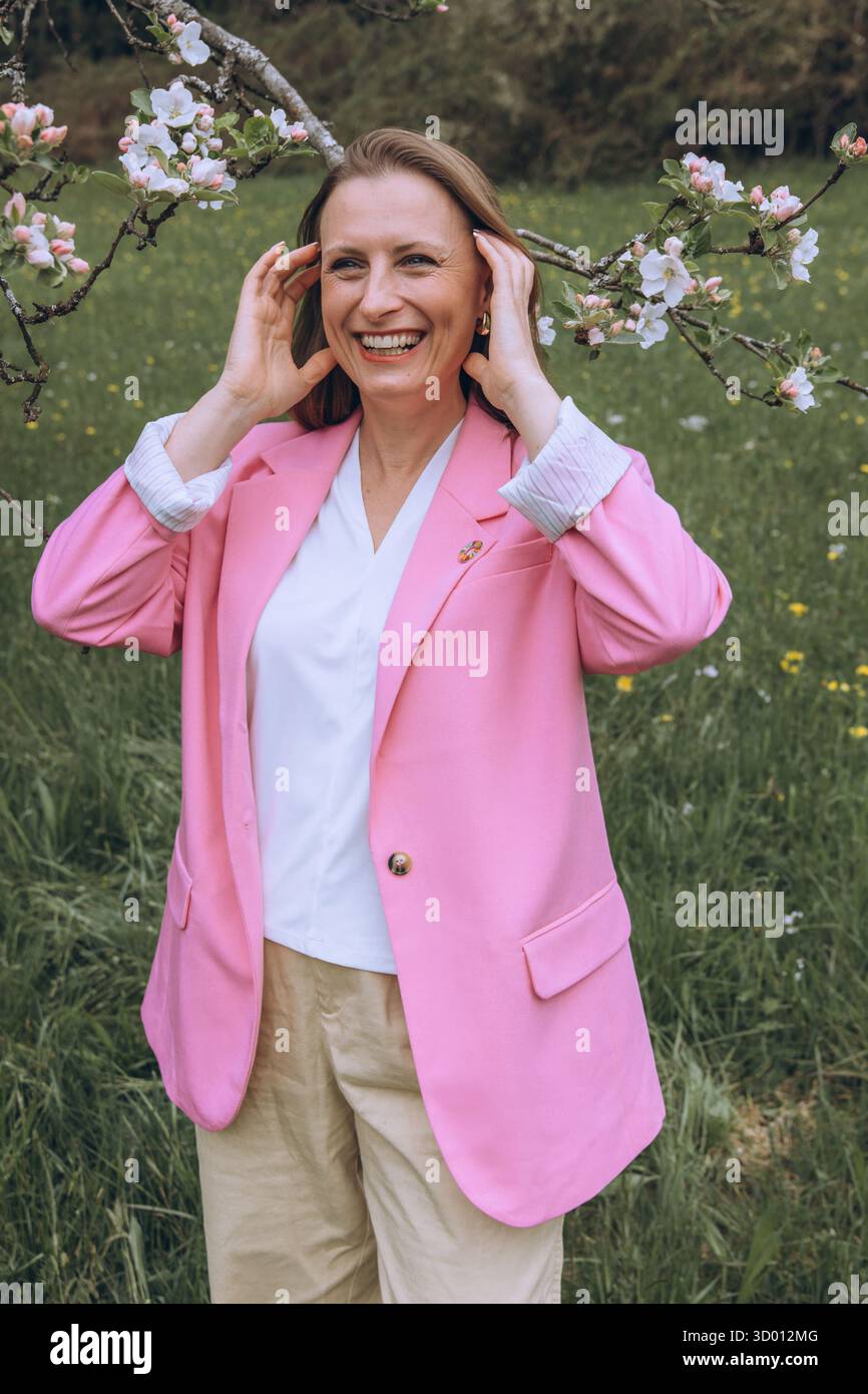 Una donna sorridente di mezza età si trova vicino a un albero in fiore nel giardino primaverile. Bellezza naturale, gioia e armonia con la natura. Rinnovamento stagionale, pace interiore e. Foto Stock