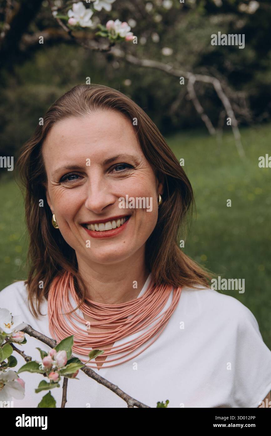 Una donna sorridente di mezza età si trova vicino a un albero in fiore nel giardino primaverile. Bellezza naturale, gioia e armonia con la natura. Rinnovamento stagionale, pace interiore e. Foto Stock