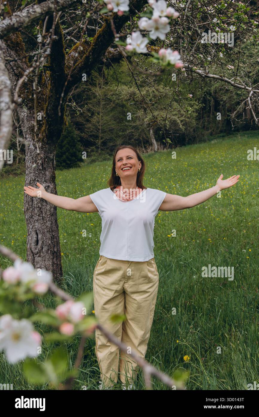 Donna sorridente di mezza età in piedi vicino a un albero in fiore nel giardino primaverile. Concetto di salute femminile, equilibrio ormonale, benessere e bellezza naturale in mi Foto Stock