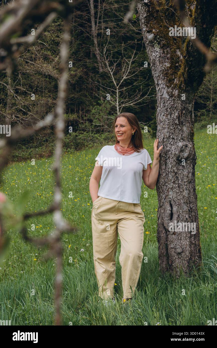 Donna sorridente di mezza età in piedi vicino a un albero in fiore nel giardino primaverile. Concetto di salute femminile, equilibrio ormonale, benessere e bellezza naturale in mi Foto Stock