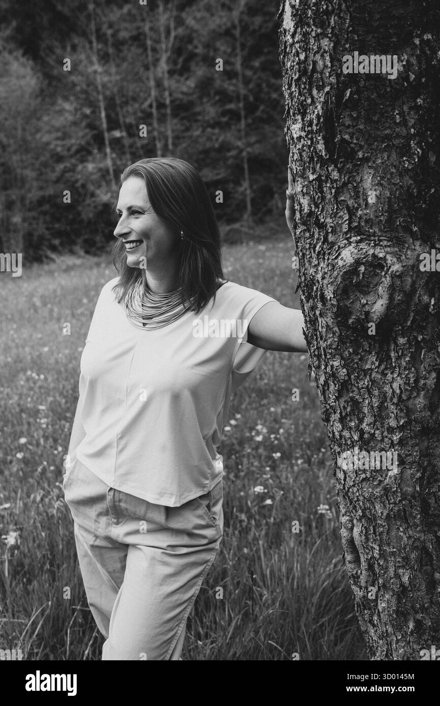 Donna sorridente di mezza età in piedi vicino a un albero in fiore nel giardino primaverile. Concetto di salute femminile, equilibrio ormonale, benessere e bellezza naturale in mi Foto Stock