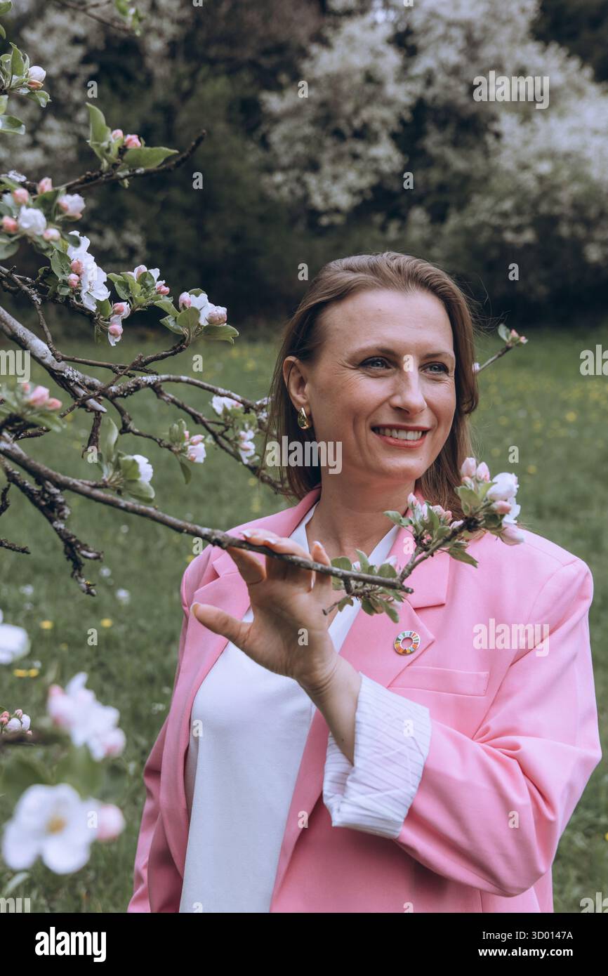 Donna sorridente di mezza età in piedi vicino a un albero in fiore nel giardino primaverile. Concetto di salute femminile, equilibrio ormonale, benessere e bellezza naturale in mi Foto Stock