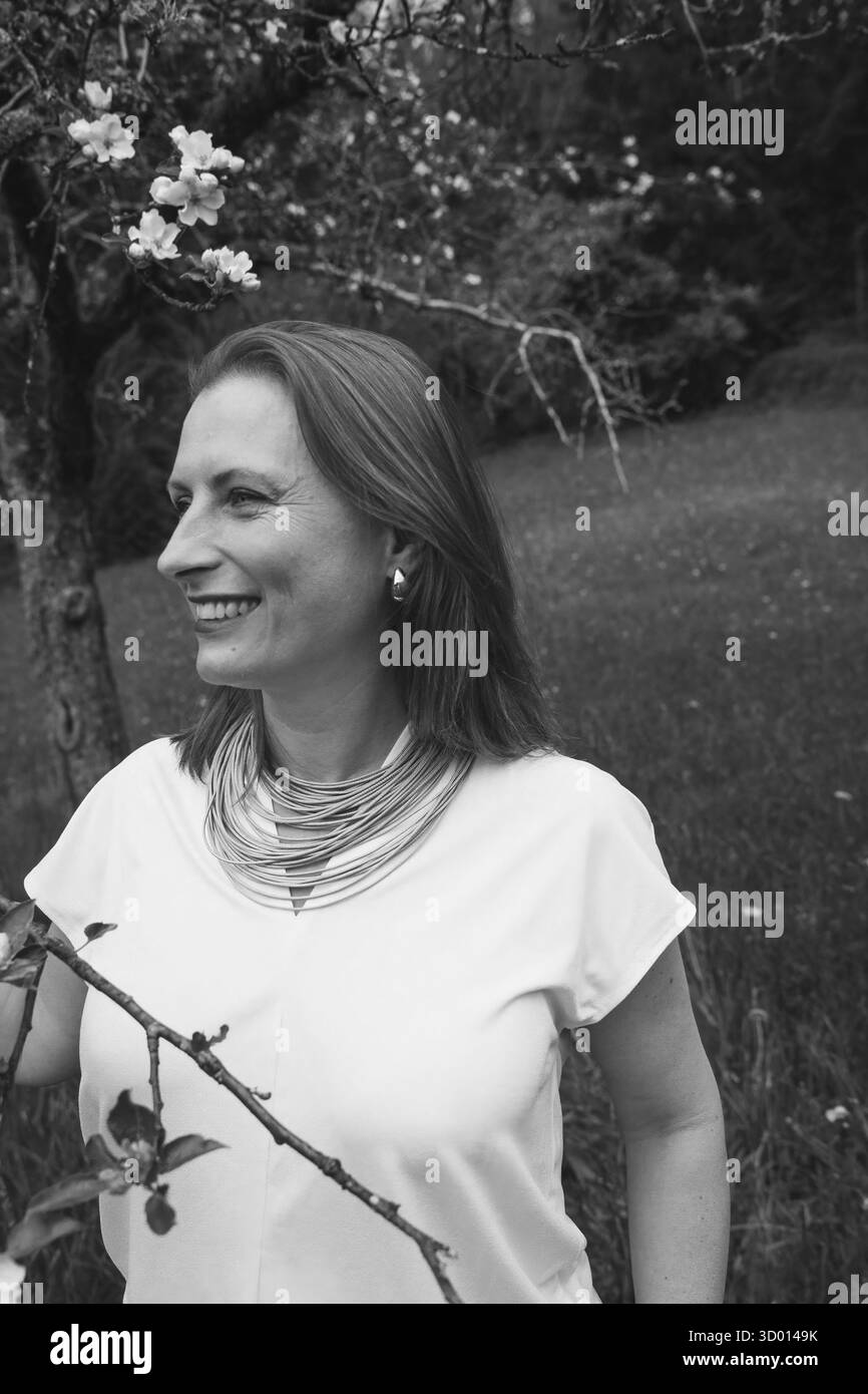 Donna sorridente di mezza età in piedi vicino a un albero in fiore nel giardino primaverile. Concetto di salute femminile, equilibrio ormonale, benessere e bellezza naturale in mi Foto Stock