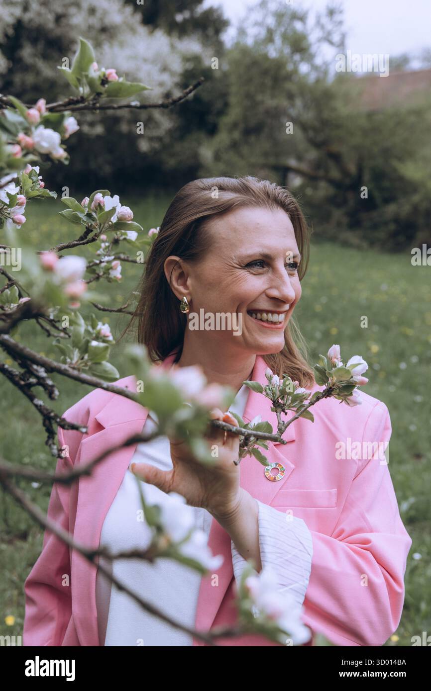 Donna sorridente di mezza età in piedi vicino a un albero in fiore nel giardino primaverile. Concetto di salute femminile, equilibrio ormonale, benessere e bellezza naturale in mi Foto Stock