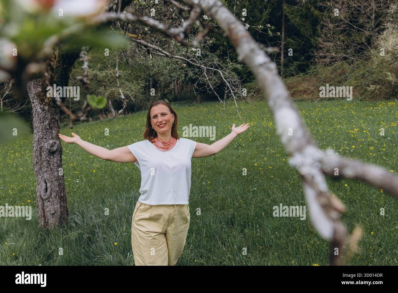 Donna sorridente di mezza età in piedi vicino a un albero in fiore nel giardino primaverile. Concetto di salute femminile, equilibrio ormonale, benessere e bellezza naturale in mi Foto Stock