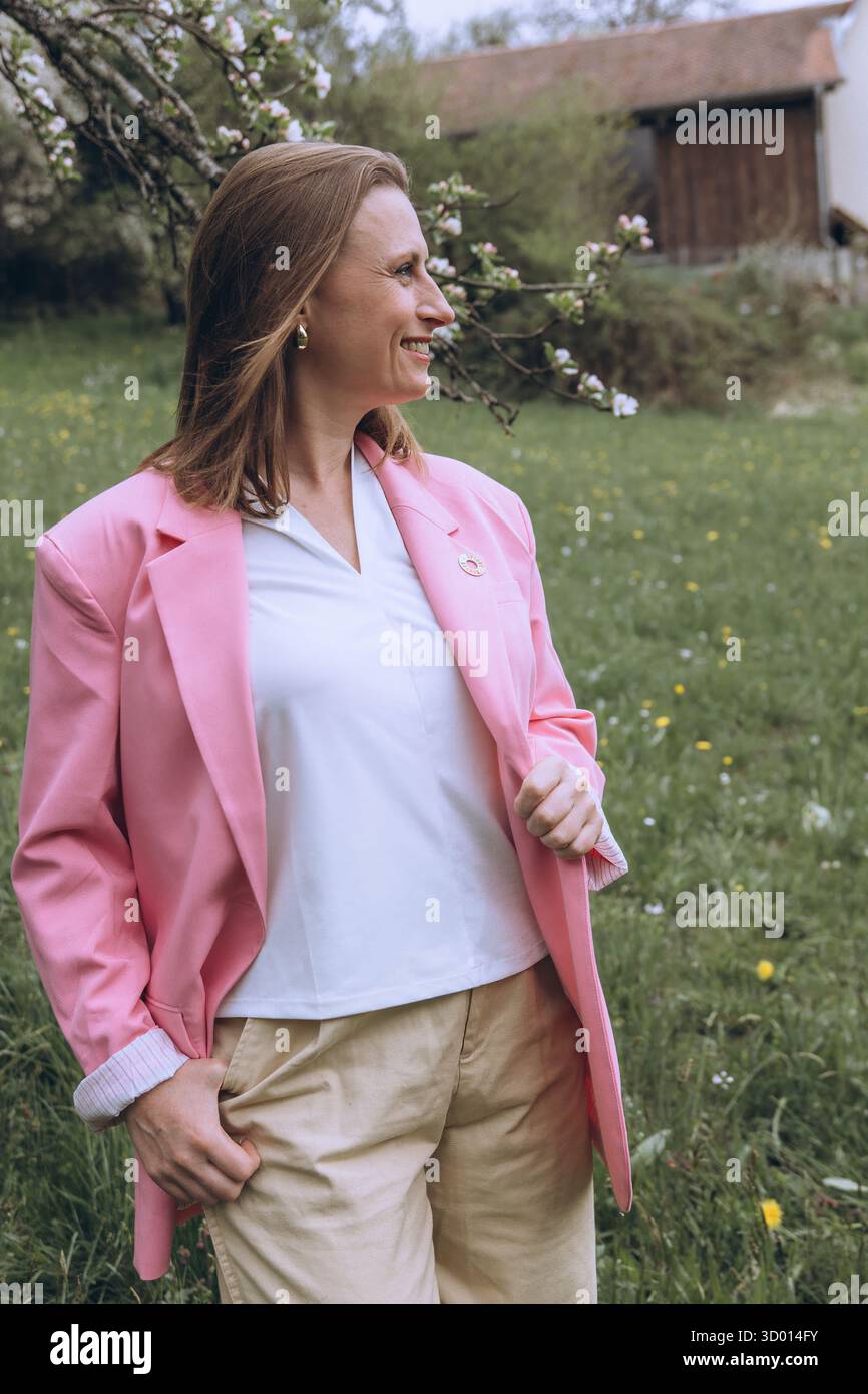 Donna sorridente di mezza età in piedi vicino a un albero in fiore nel giardino primaverile. Concetto di salute femminile, equilibrio ormonale, benessere e bellezza naturale in mi Foto Stock