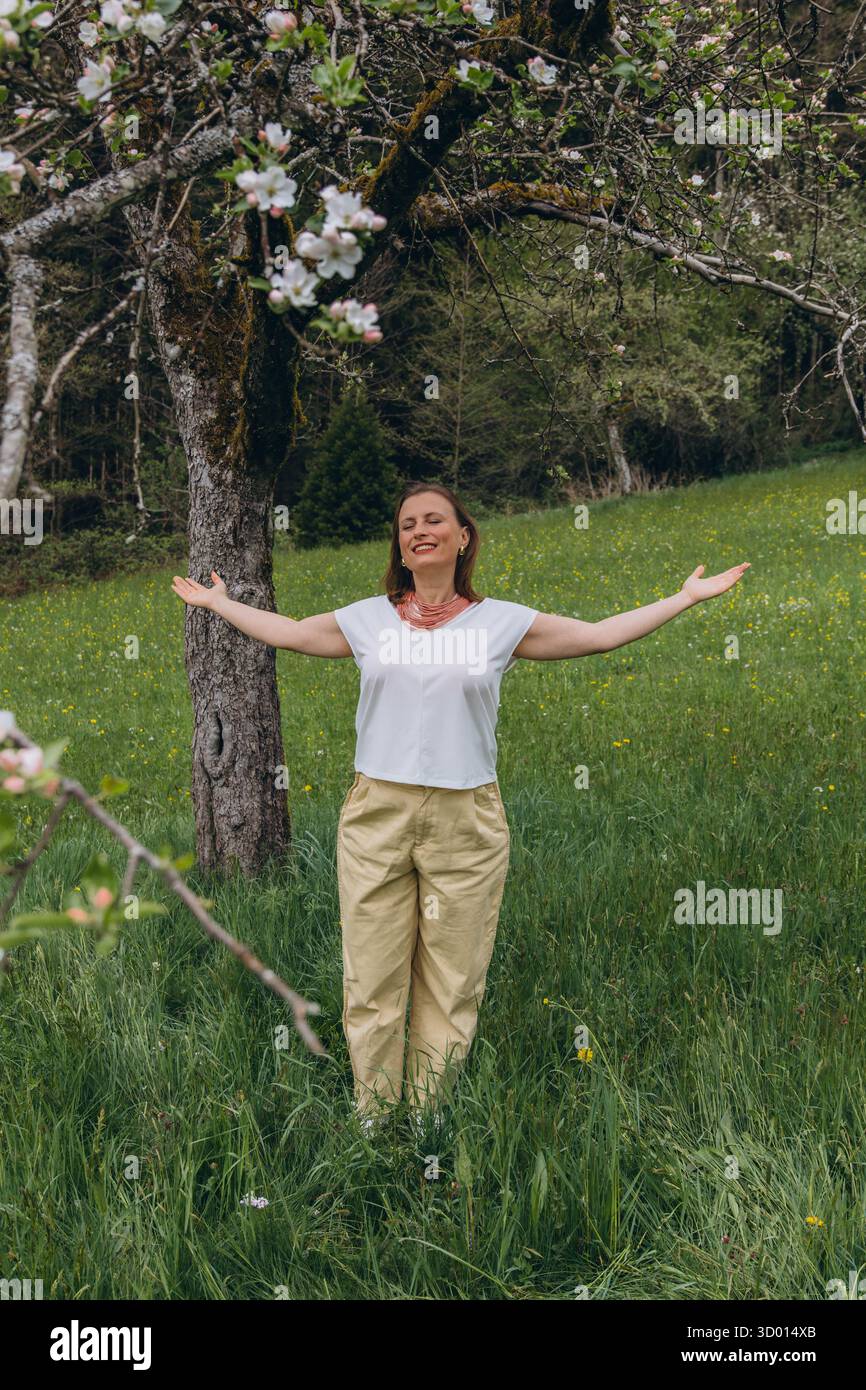 Donna sorridente di mezza età in piedi vicino a un albero in fiore nel giardino primaverile. Concetto di salute femminile, equilibrio ormonale, benessere e bellezza naturale in mi Foto Stock