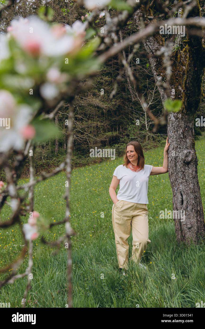 Donna sorridente di mezza età in piedi vicino a un albero in fiore nel giardino primaverile. Concetto di salute femminile, equilibrio ormonale, benessere e bellezza naturale in mi Foto Stock