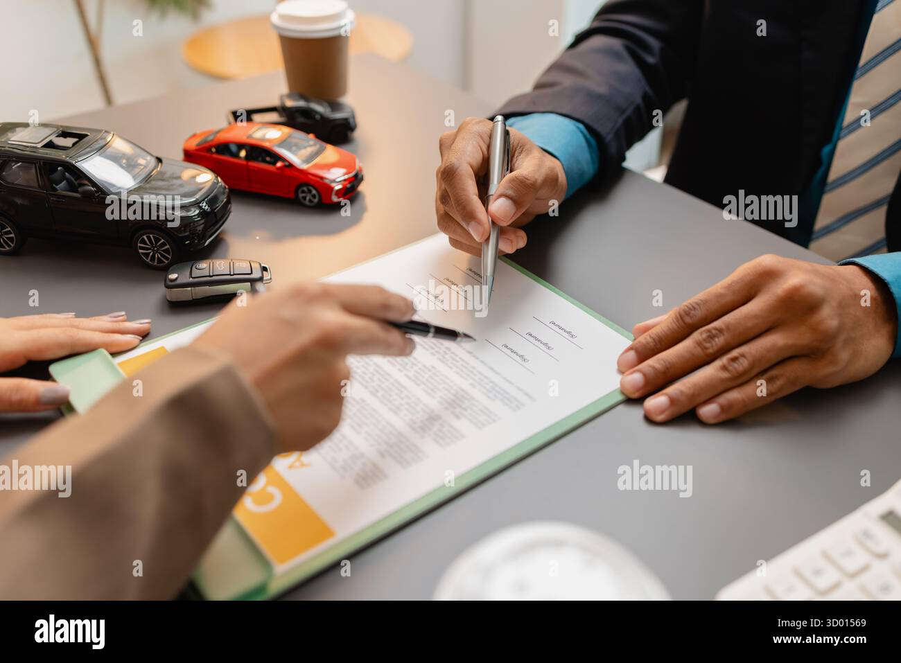 Un uomo d'affari e un cliente stanno esaminando e firmando un contratto di vendita di auto presso una scrivania, con modellini di auto di piccole dimensioni, una tazza di caffè e documenti intorno a loro, Foto Stock