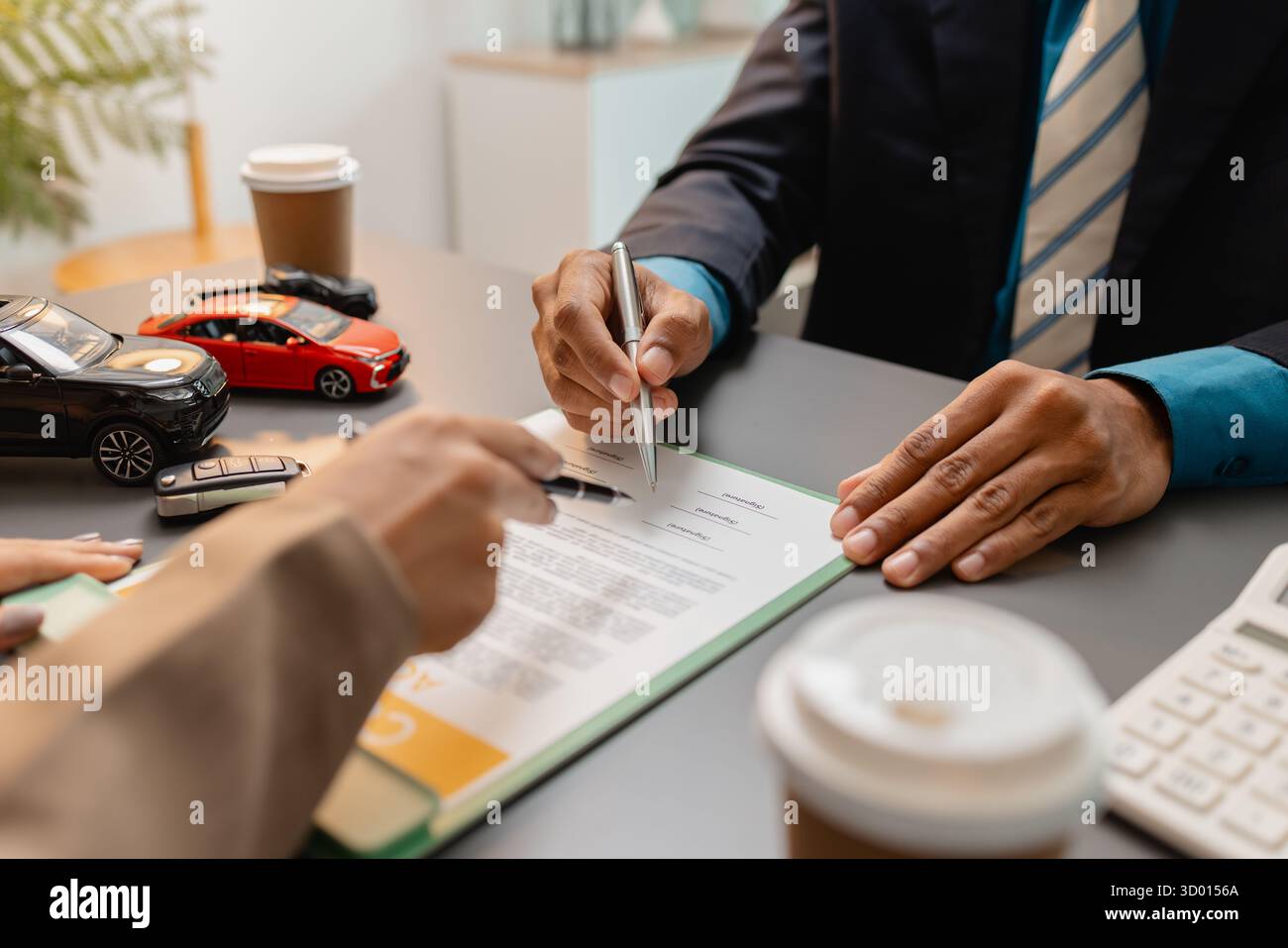 Un uomo d'affari e un cliente stanno esaminando e firmando un contratto di vendita di auto presso una scrivania, con modellini di auto di piccole dimensioni, una tazza di caffè e documenti intorno a loro, Foto Stock