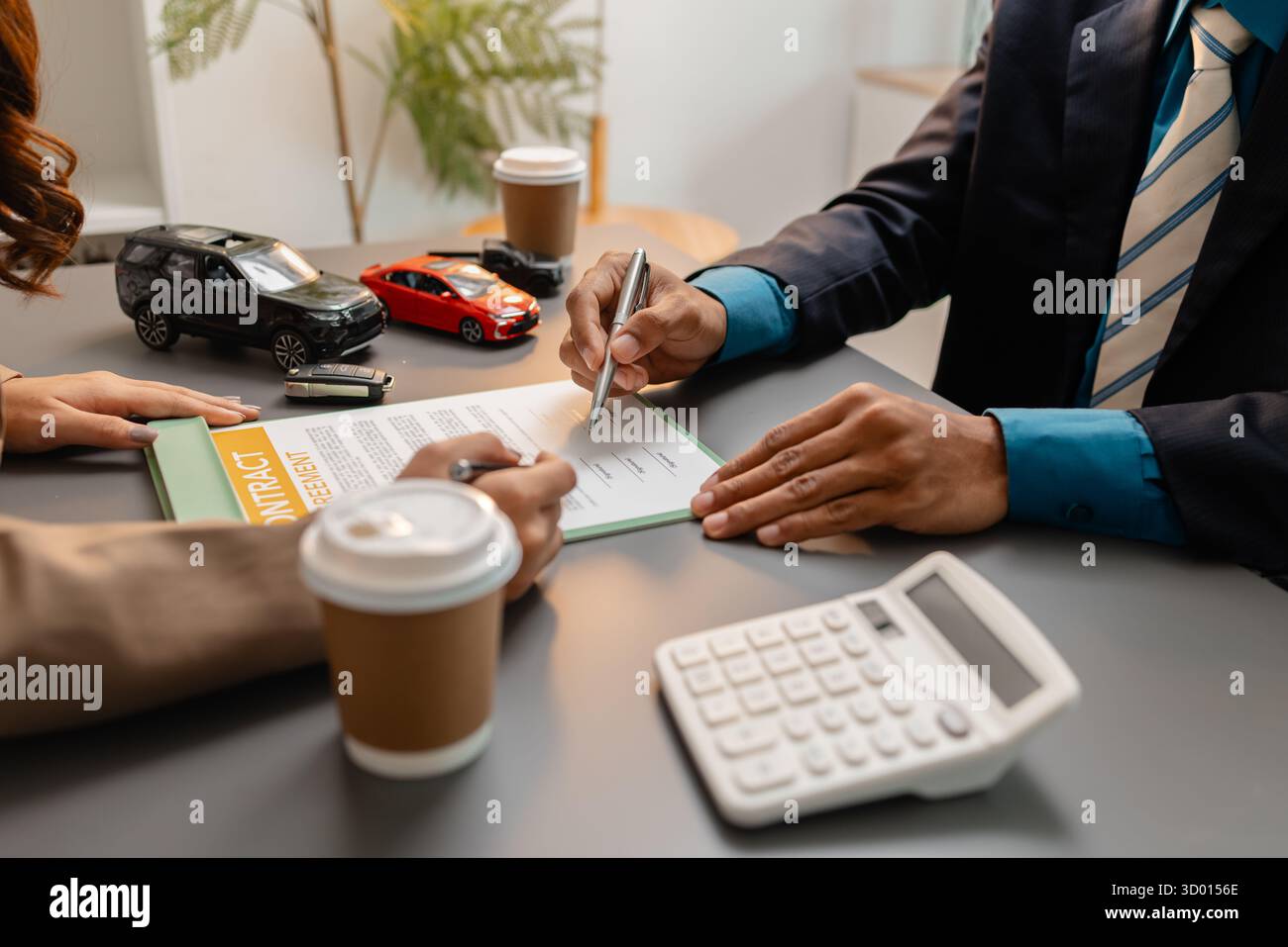 Un uomo d'affari e un cliente stanno esaminando e firmando un contratto di vendita di auto presso una scrivania, con modellini di auto di piccole dimensioni, una tazza di caffè e documenti intorno a loro, Foto Stock