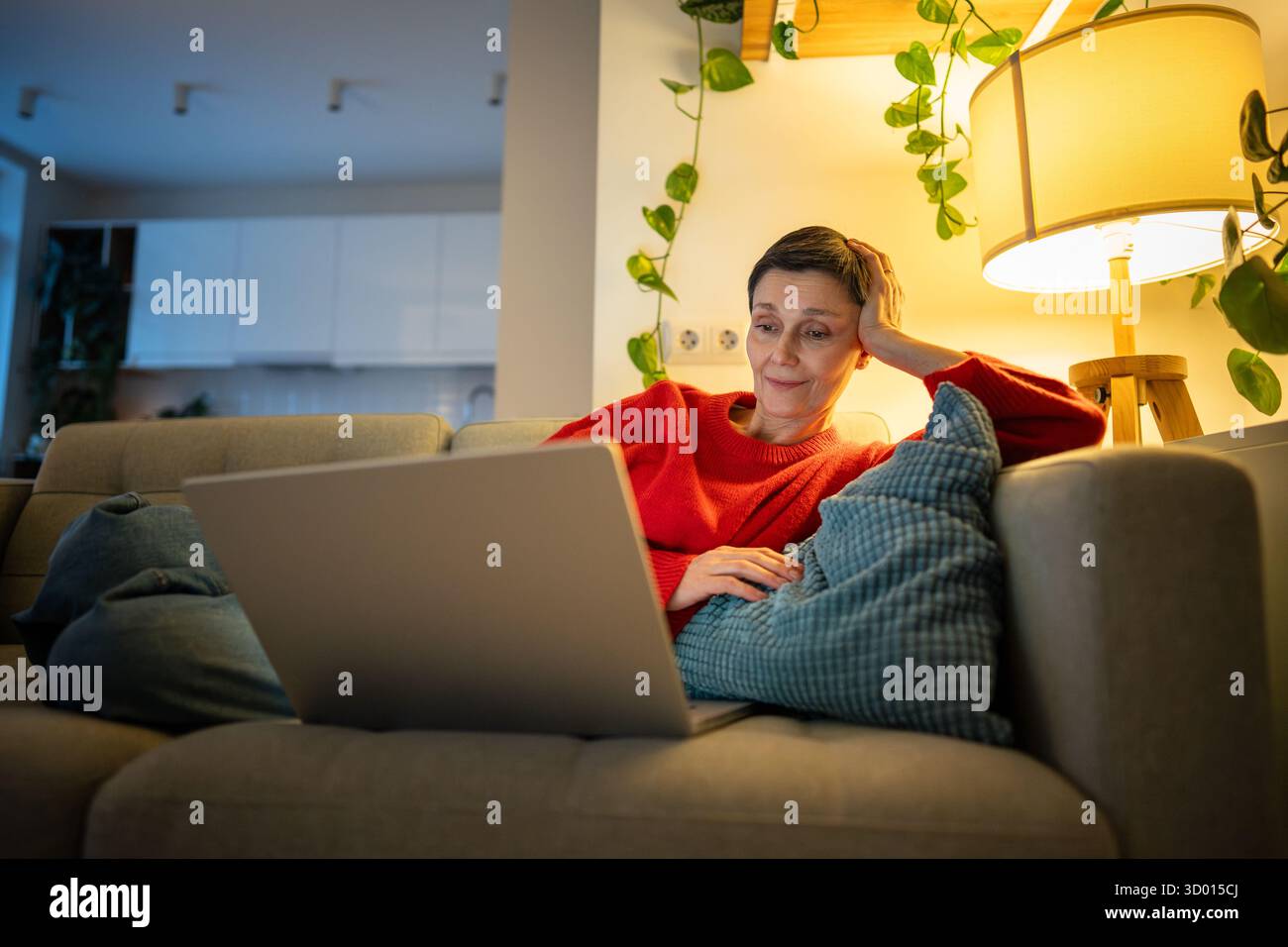 Una donna sorridente e felice sdraiata sul divano guardando film sul laptop in un salotto accogliente da sola, riposando le donne Foto Stock Una donna sorridente e felice sdraiata sul divano guardando film sul laptop in un salotto accogliente da sola, riposando le donne Foto Stock